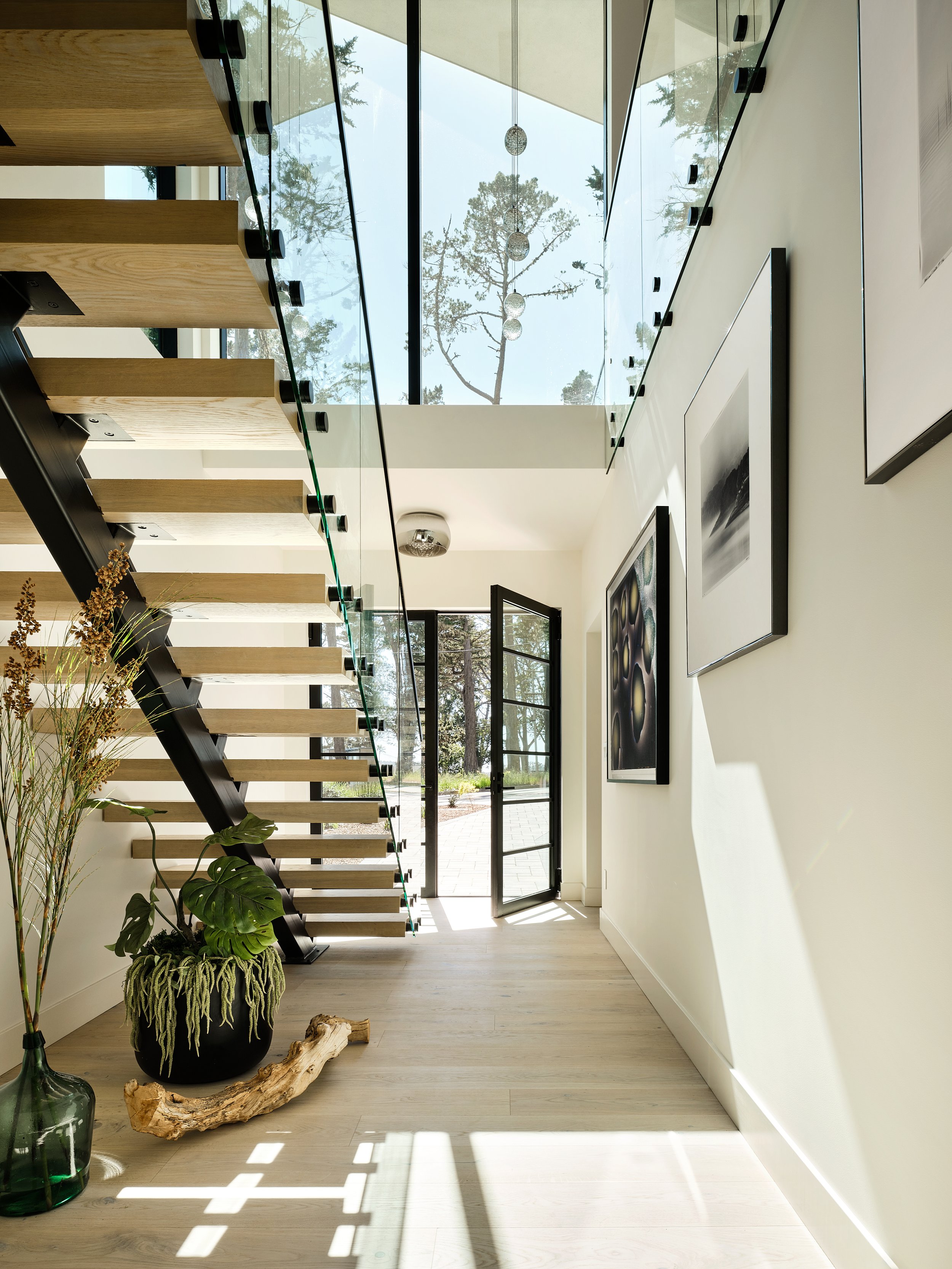 Interior of a modern house featuring a floating staircase with wooden steps and glass railings, sunlight streaming through large glass doors and windows, with framed art on white walls and plants near the entrance.