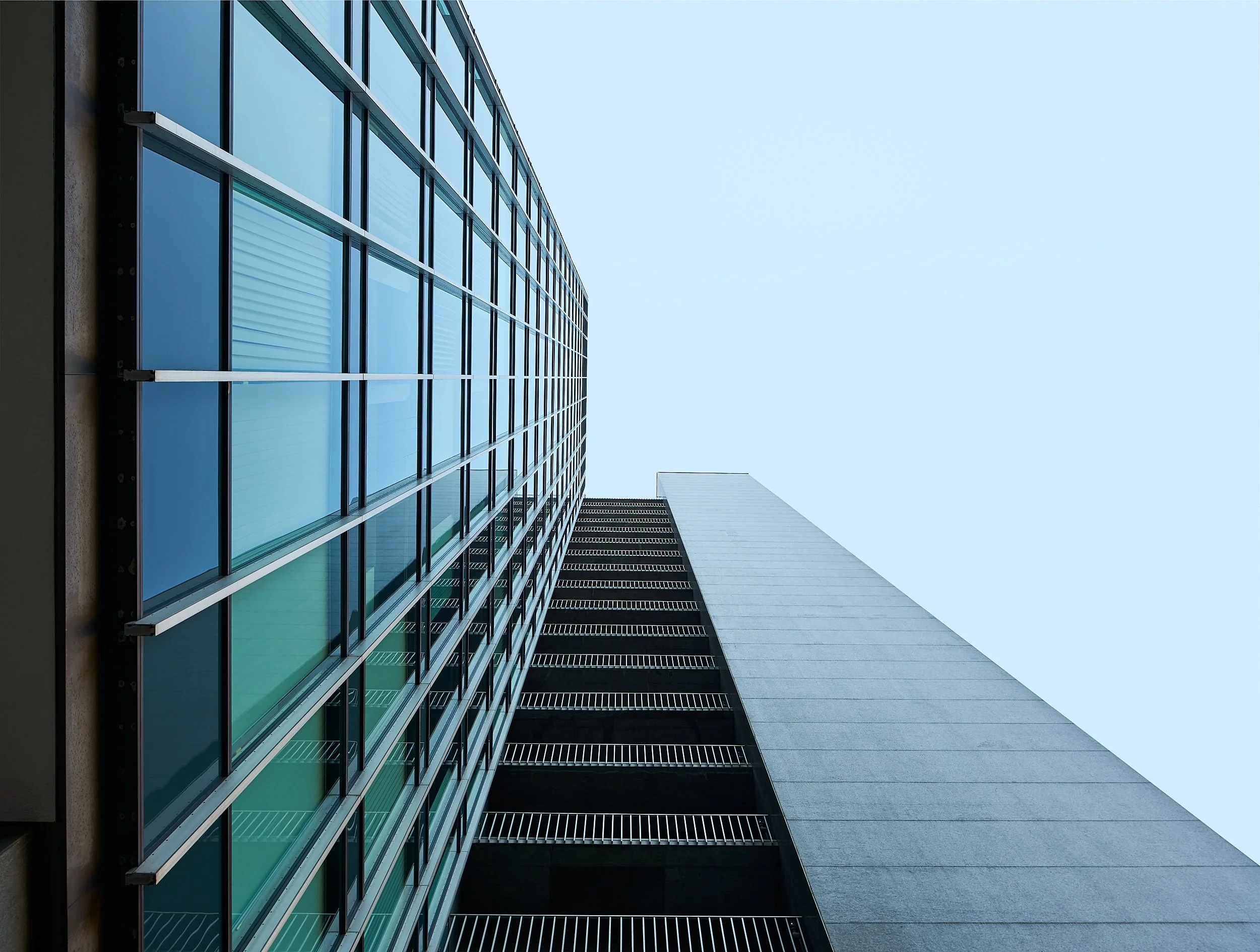 Low-angle view of modern high-rise building with glass windows and concrete structure against a clear blue sky.