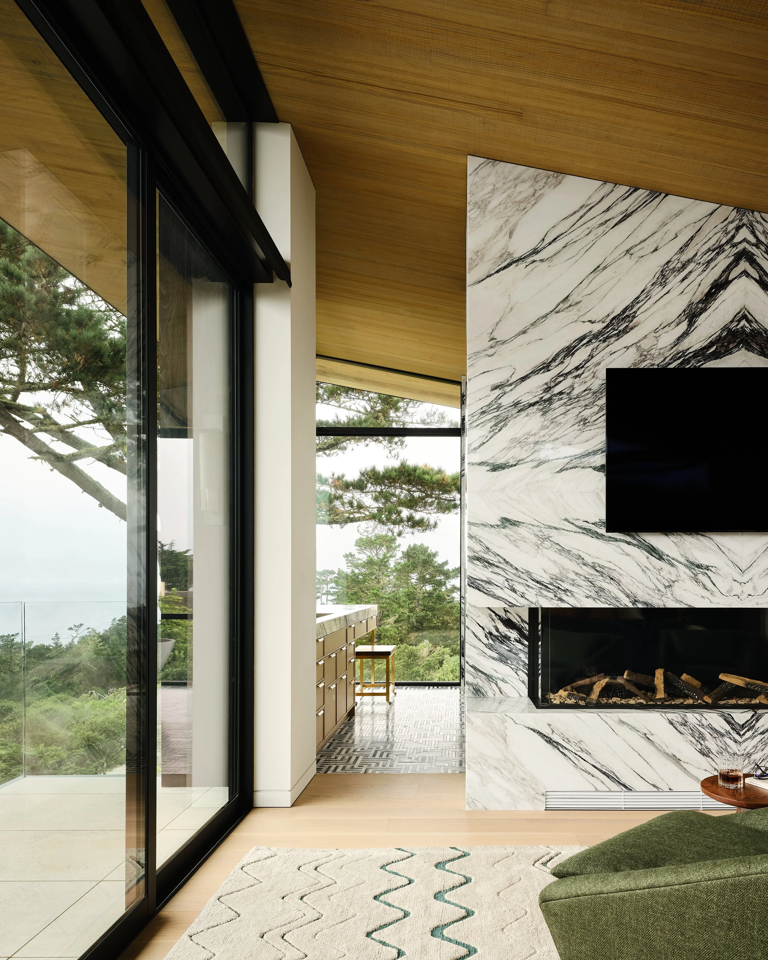 Modern living room with a marble fireplace, mounted flat-screen TV, large glass sliding door, wooden ceiling, and outdoor view of trees.