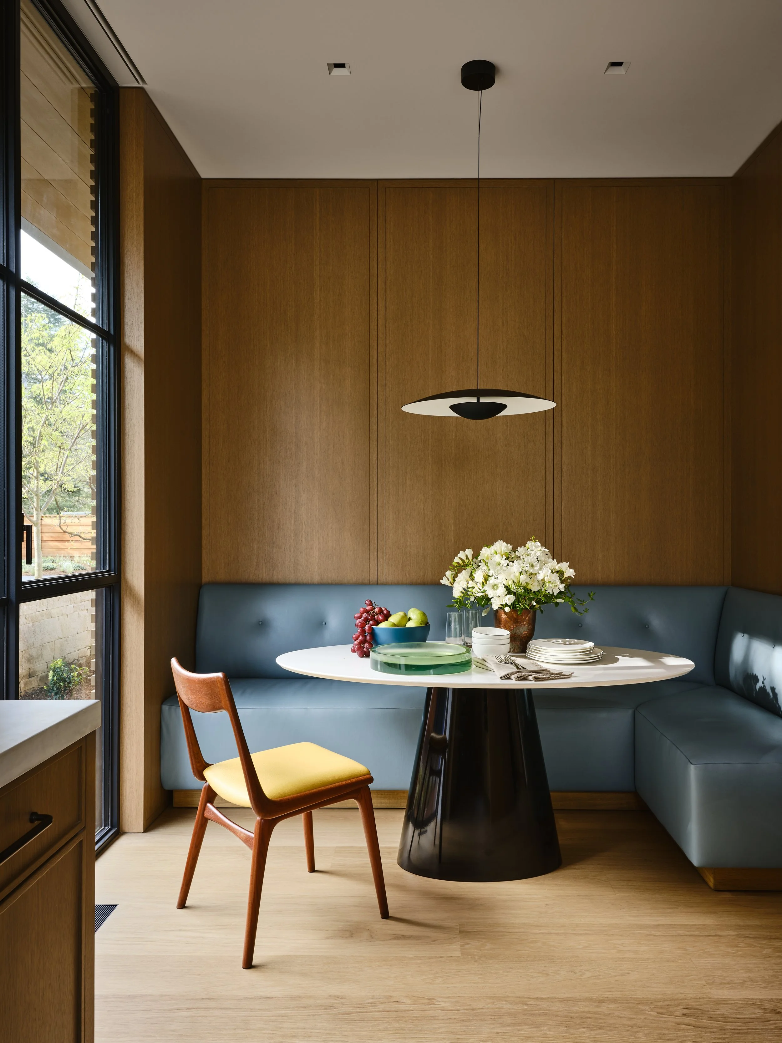 Modern dining nook with a round white table, blue built-in bench seating, a single wooden chair with yellow cushion, a vase of white flowers, grapes, apples, and tableware, with a large window and wood-paneled wall. Interior design and Architecture. 