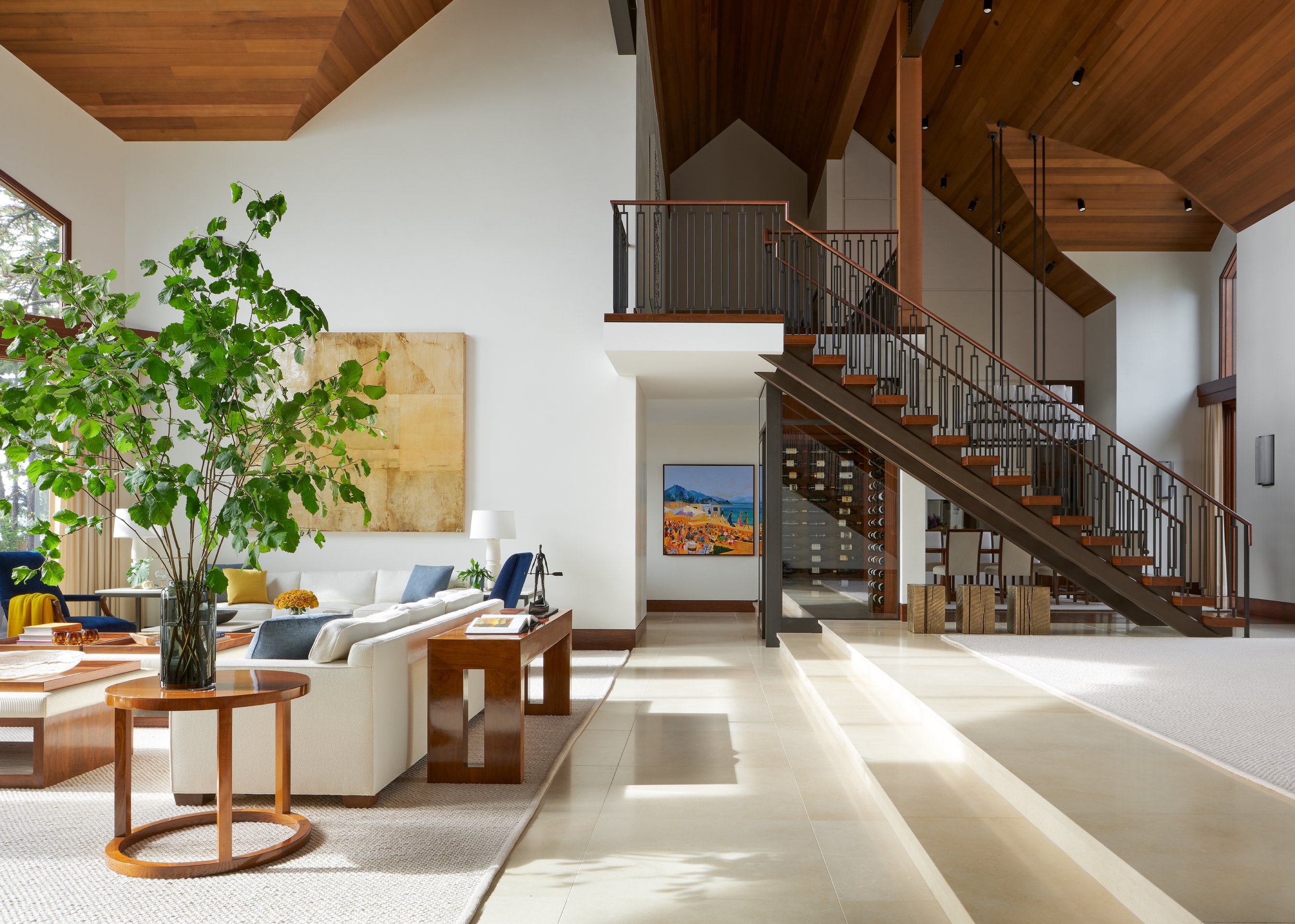 Bright living room with a large green plant, white sofas, wooden furniture, and an open staircase with wooden steps and black metal railings, high vaulted ceilings, and artwork on the walls.