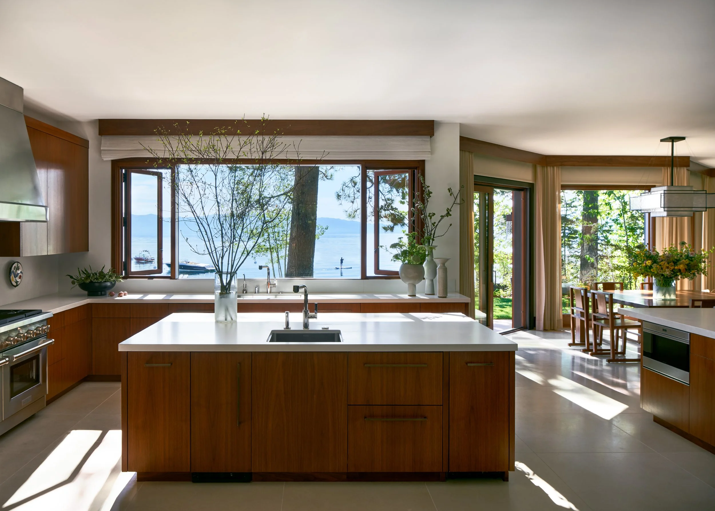 Modern kitchen with wooden cabinets and a large island, overlooking a lake through wide windows adorned with plants and flowers, with natural sunlight illuminating the space.