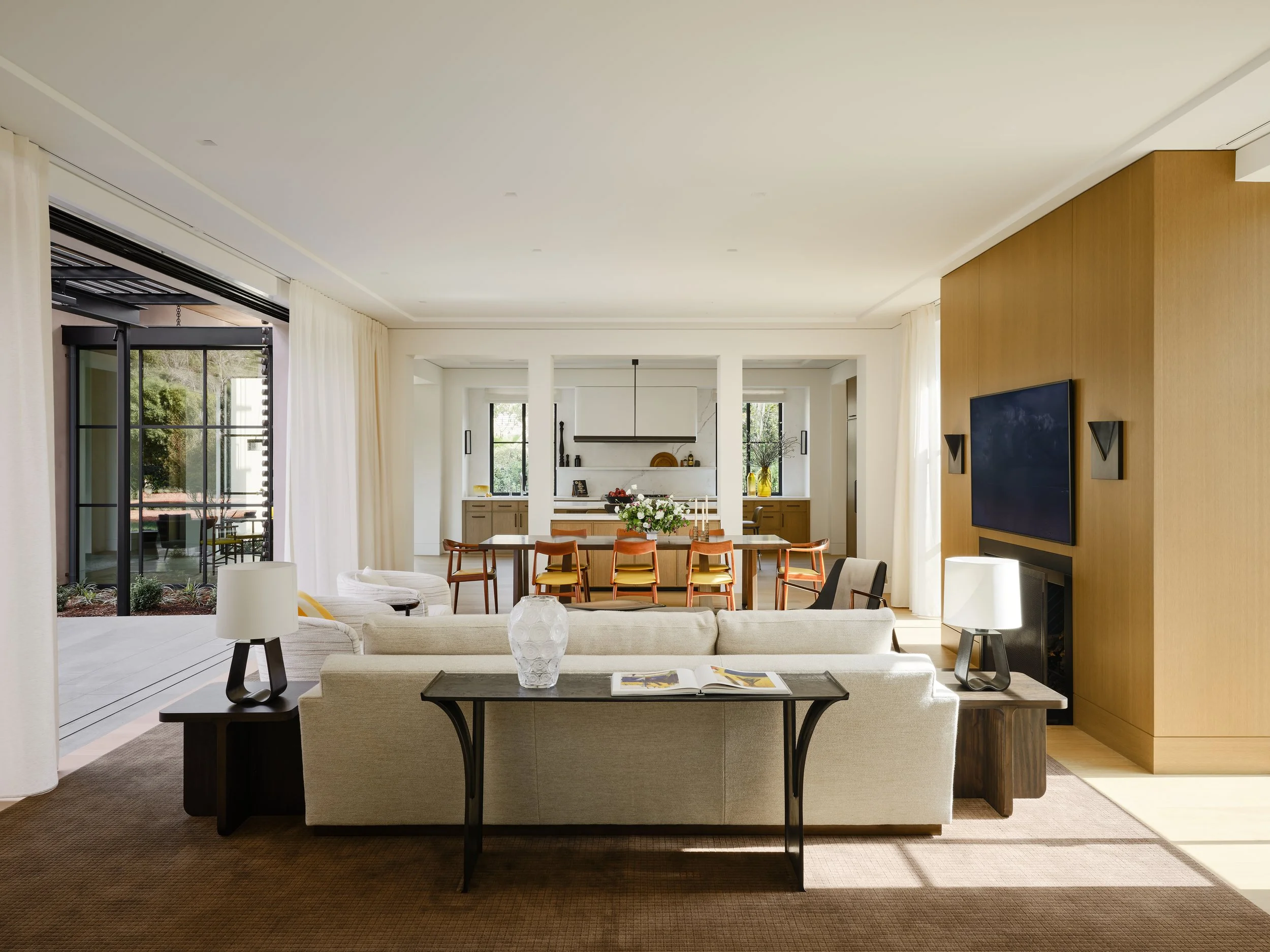 Living room with beige sofa, black side tables with lamps, a white vase, and open book, facing a wall-mounted TV on a wooden accent wall. Behind the sofa is a dining area with a table and multiple chairs, and kitchen with windows and cabinets in the 