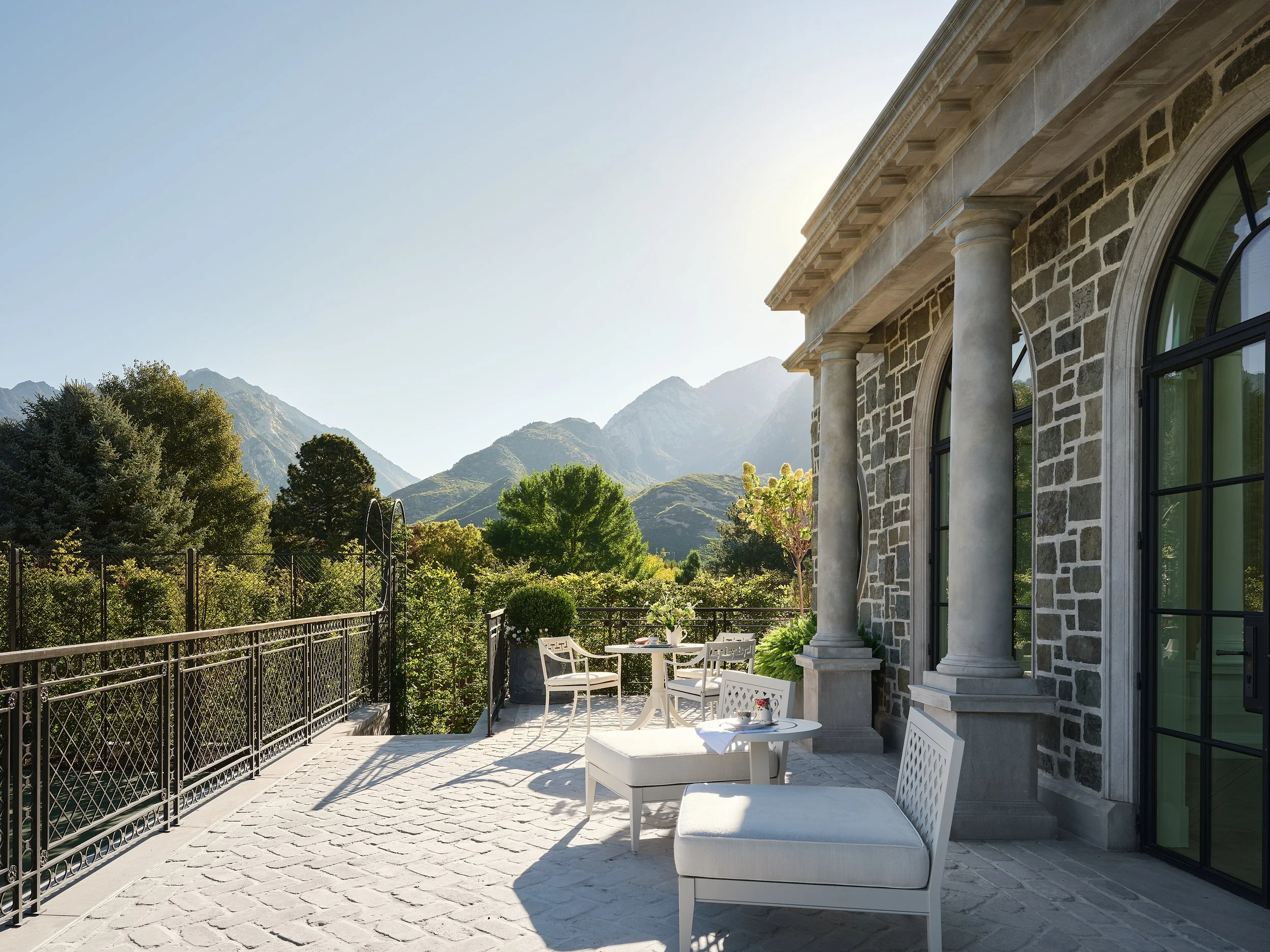 A spacious outdoor patio with white outdoor furniture, including chairs and a table, overlooking a lush green landscape and mountains in the background. The patio is attached to a stone building with large arched windows and classical columns.