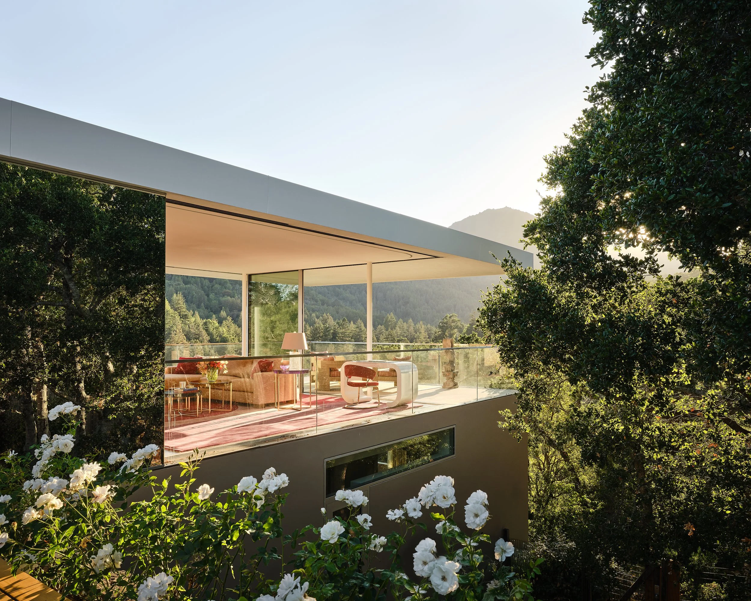 Modern house with large glass windows, an outdoor balcony overlooking a lush green landscape, trees with white flowers in the foreground, mountains in the background, and sunlight shining through the trees.