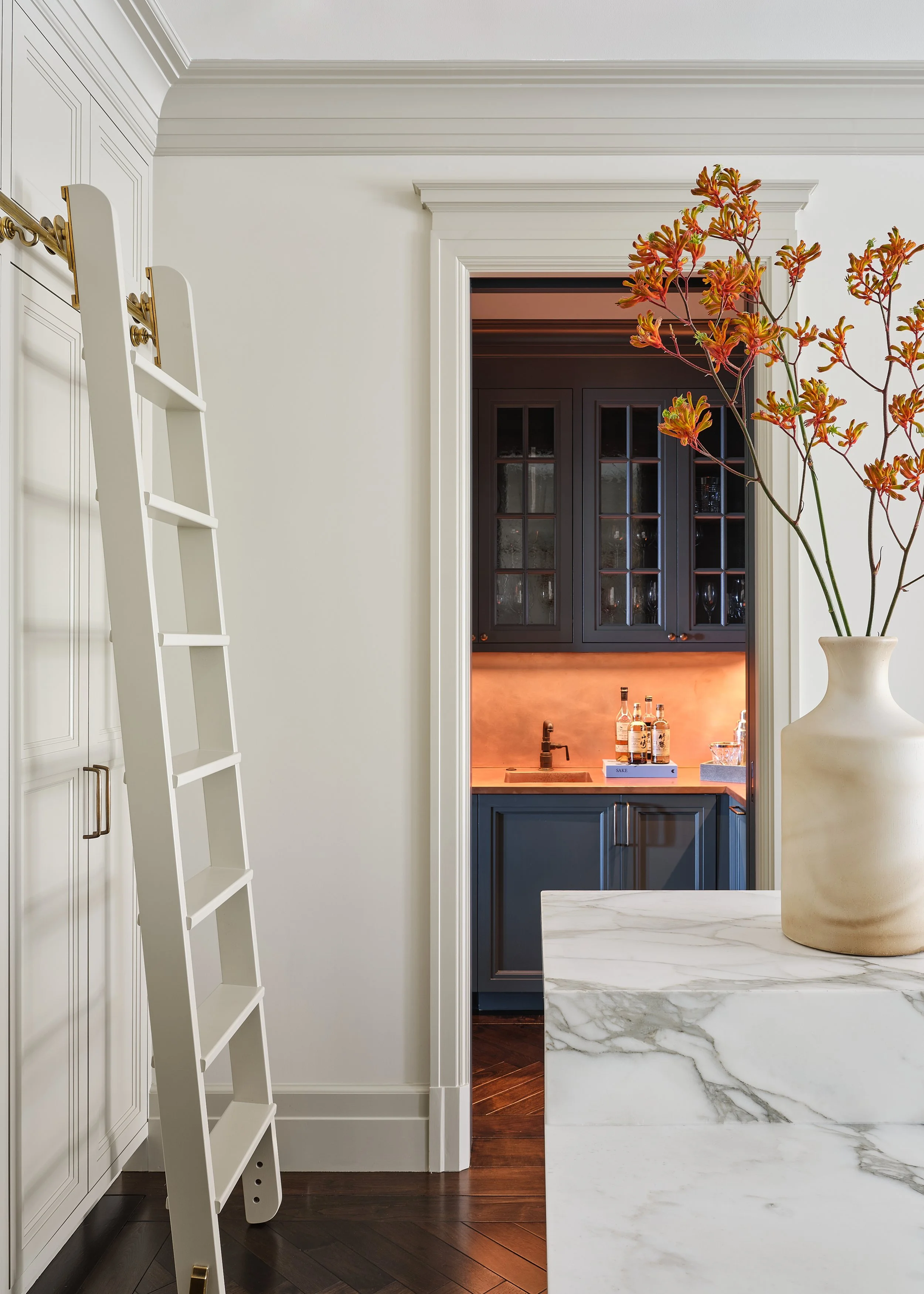 Interior view of a modern home with a kitchen visible through a doorway. The foreground features a large white marble table topped with a beige vase holding orange and red flowers. To the left, there is a white ladder leaning against a wall, and to t