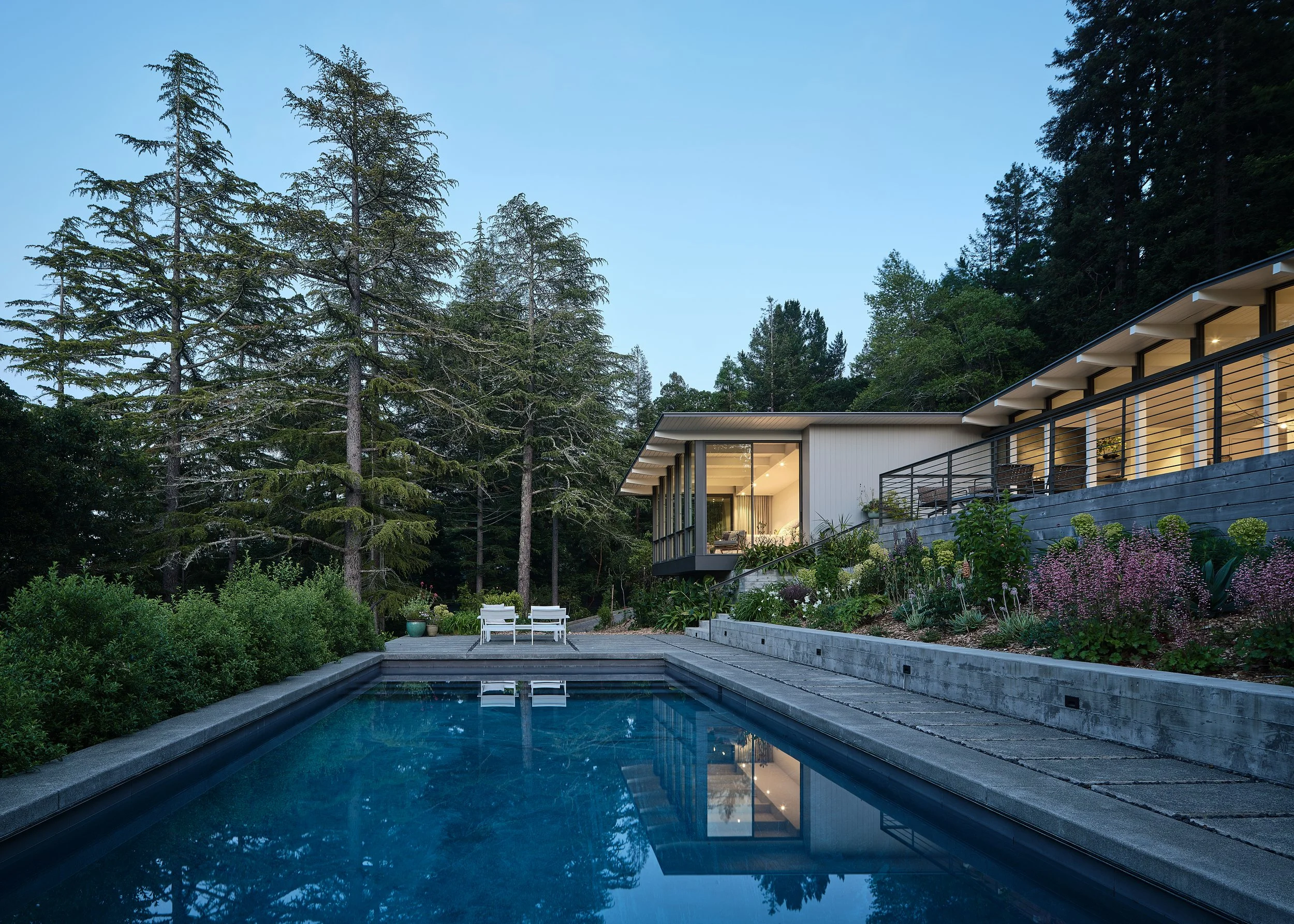 A modern house with large glass windows and a balcony sits in a wooded area, with a backyard featuring a swimming pool and garden beds with blooming flowers.