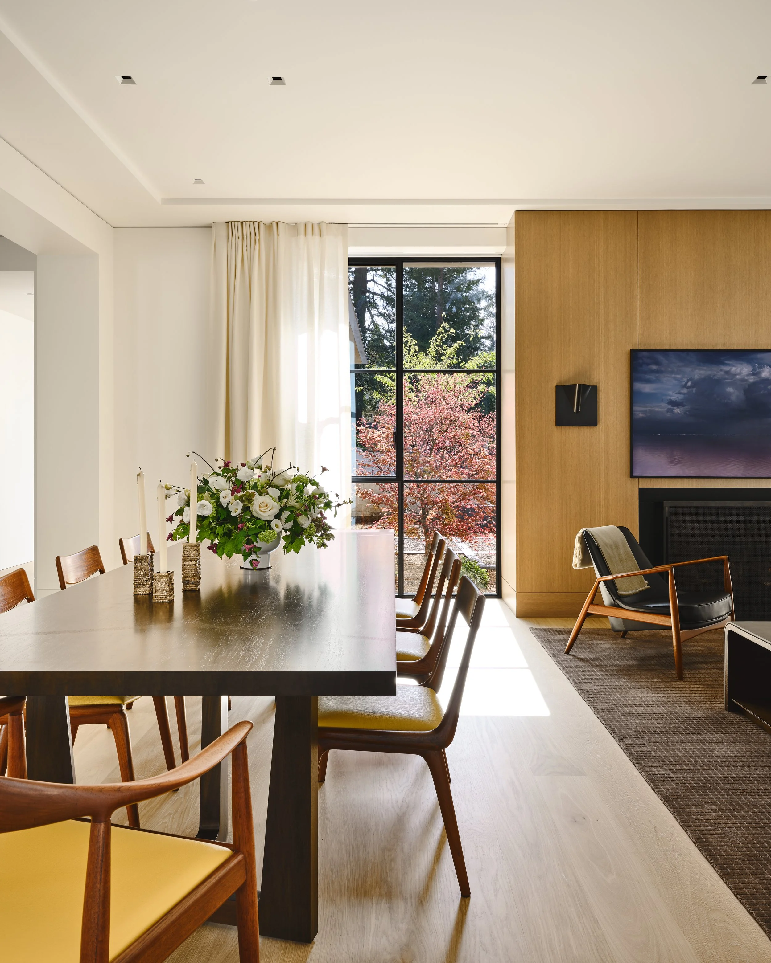 Modern dining room with a wooden table, yellow chairs, a flower centerpiece, and a large window showing trees outside, with sunlight streaming in. Interior design and Architecture. California architectural photographer
