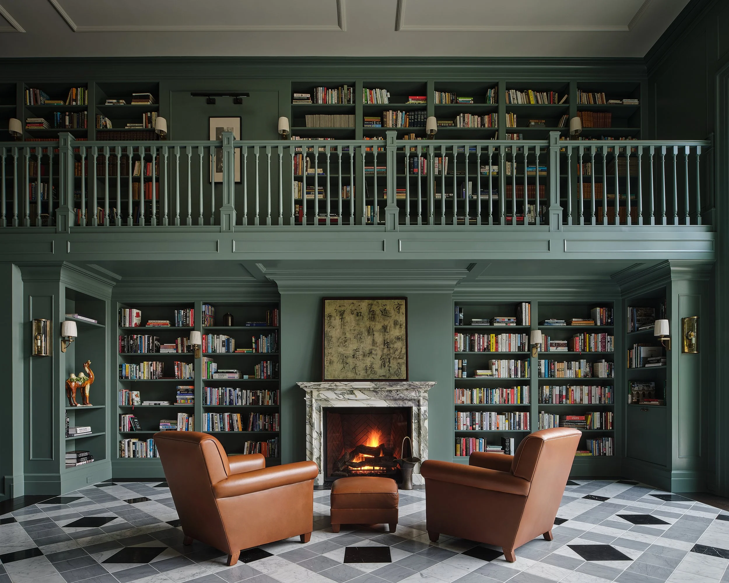 A cozy living room with a fireplace, surrounded by built-in bookshelves filled with books, two brown leather armchairs and a small matching ottoman in front of the fireplace.