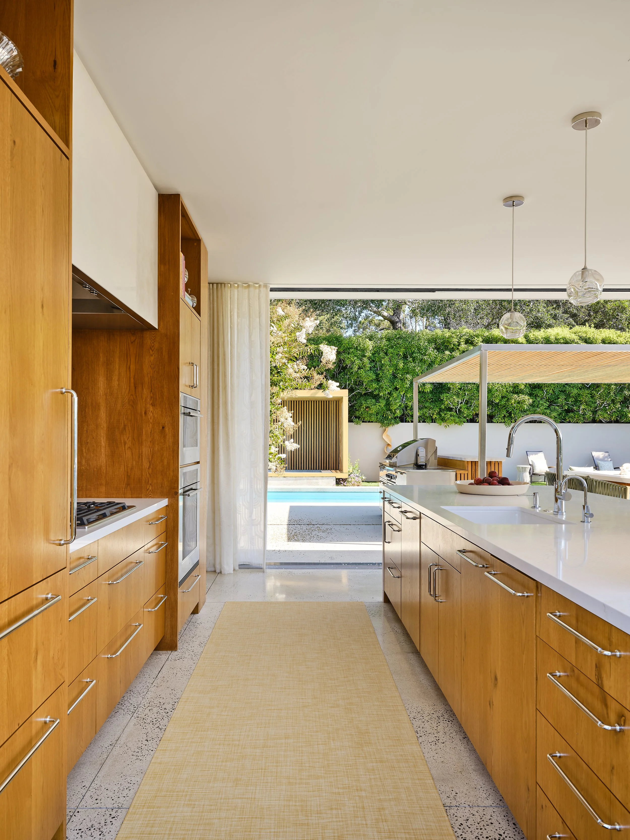 Modern kitchen with wooden cabinets, white countertops, and a view of a backyard with a pool, greenery, and outdoor seating.