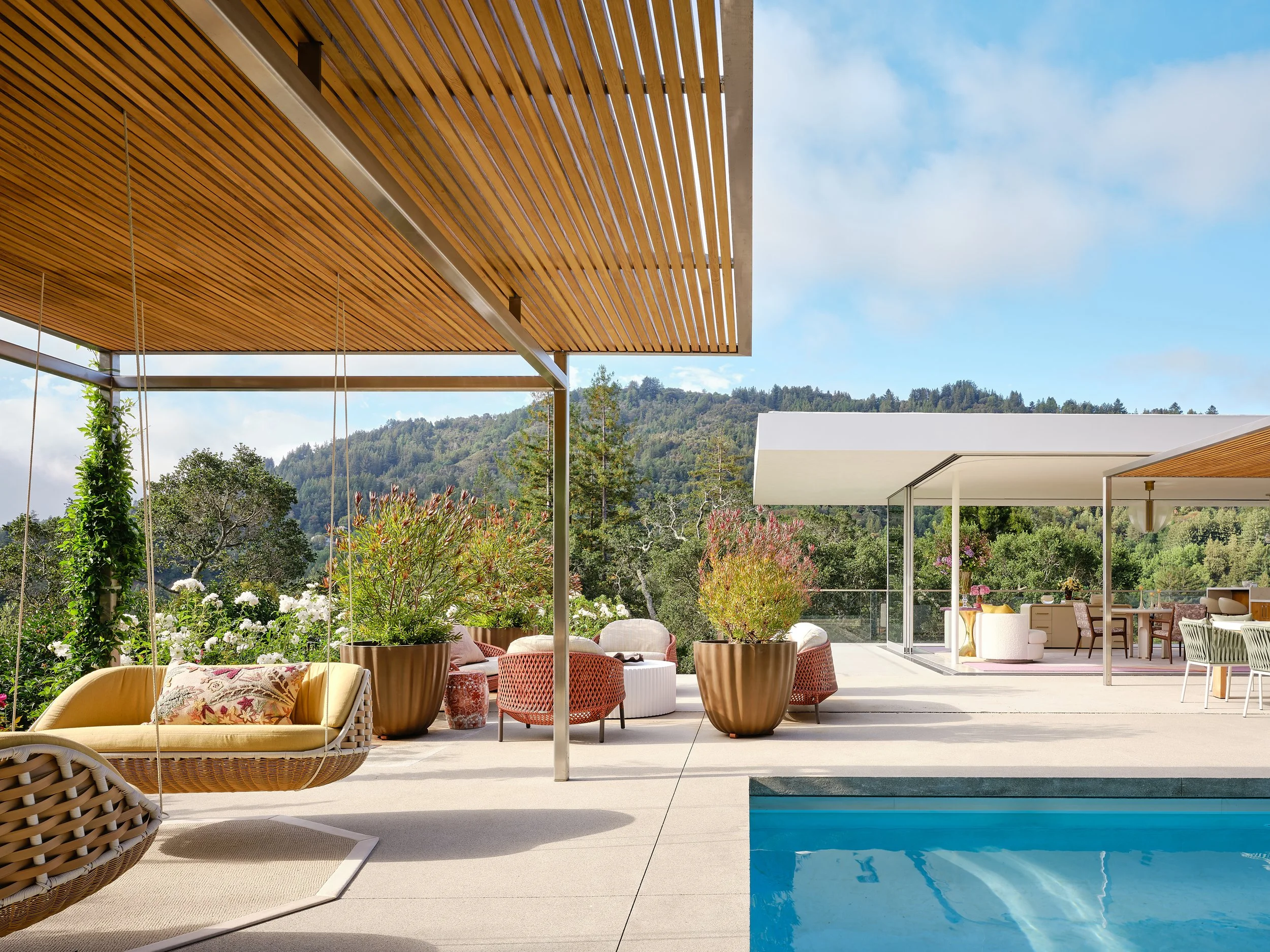 Modern outdoor patio area with seating, planters, and a swimming pool with forested hills in the background under a partly cloudy sky.