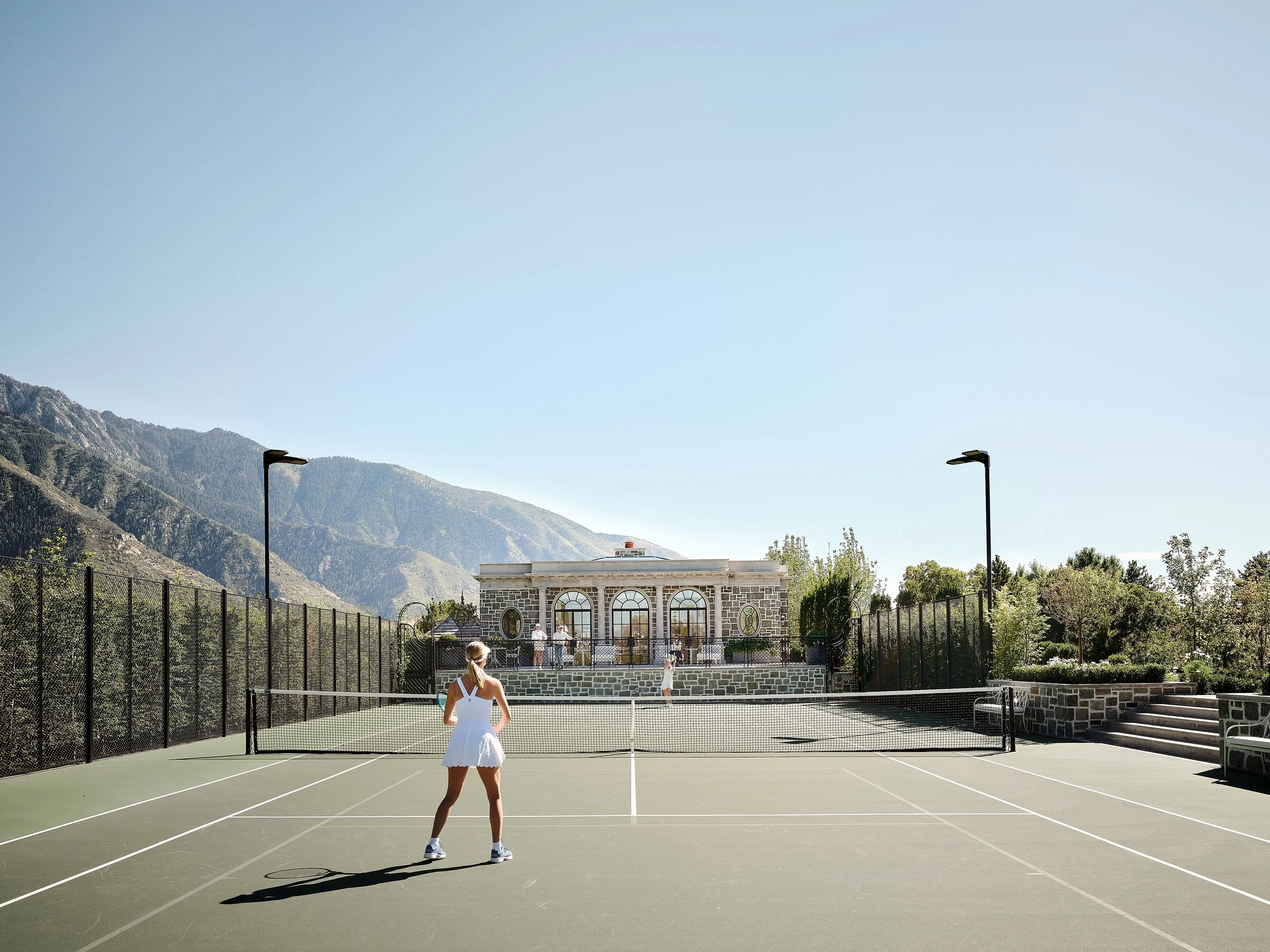A woman in a white tennis outfit standing on a green tennis court with a net, surrounded by a black fence, set against mountains and a clear blue sky.