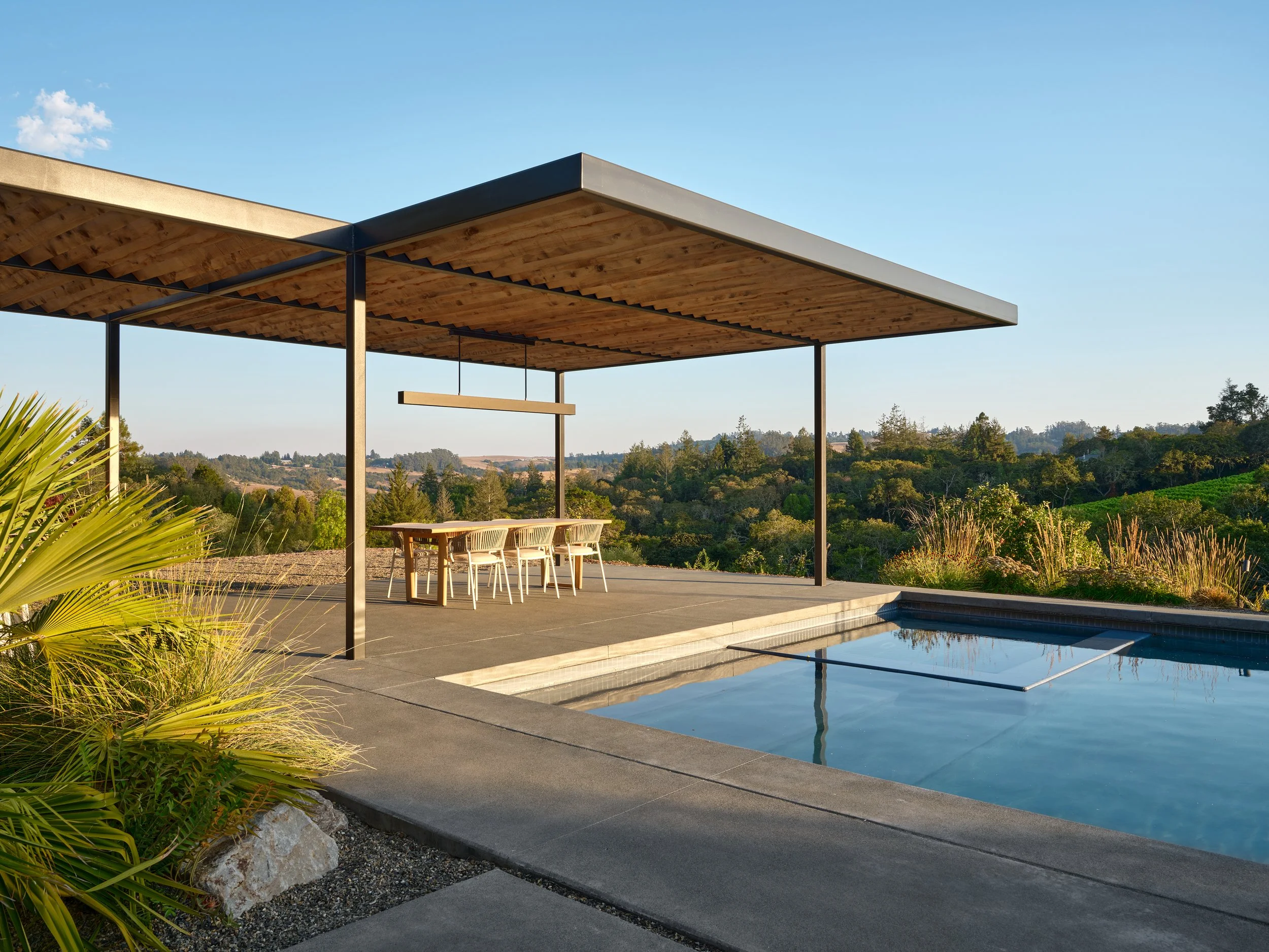 Outdoor patio with a swimming pool, modern wooden and metal pergola, dining table with six chairs, overlooking green hills and trees, under a clear blue sky.