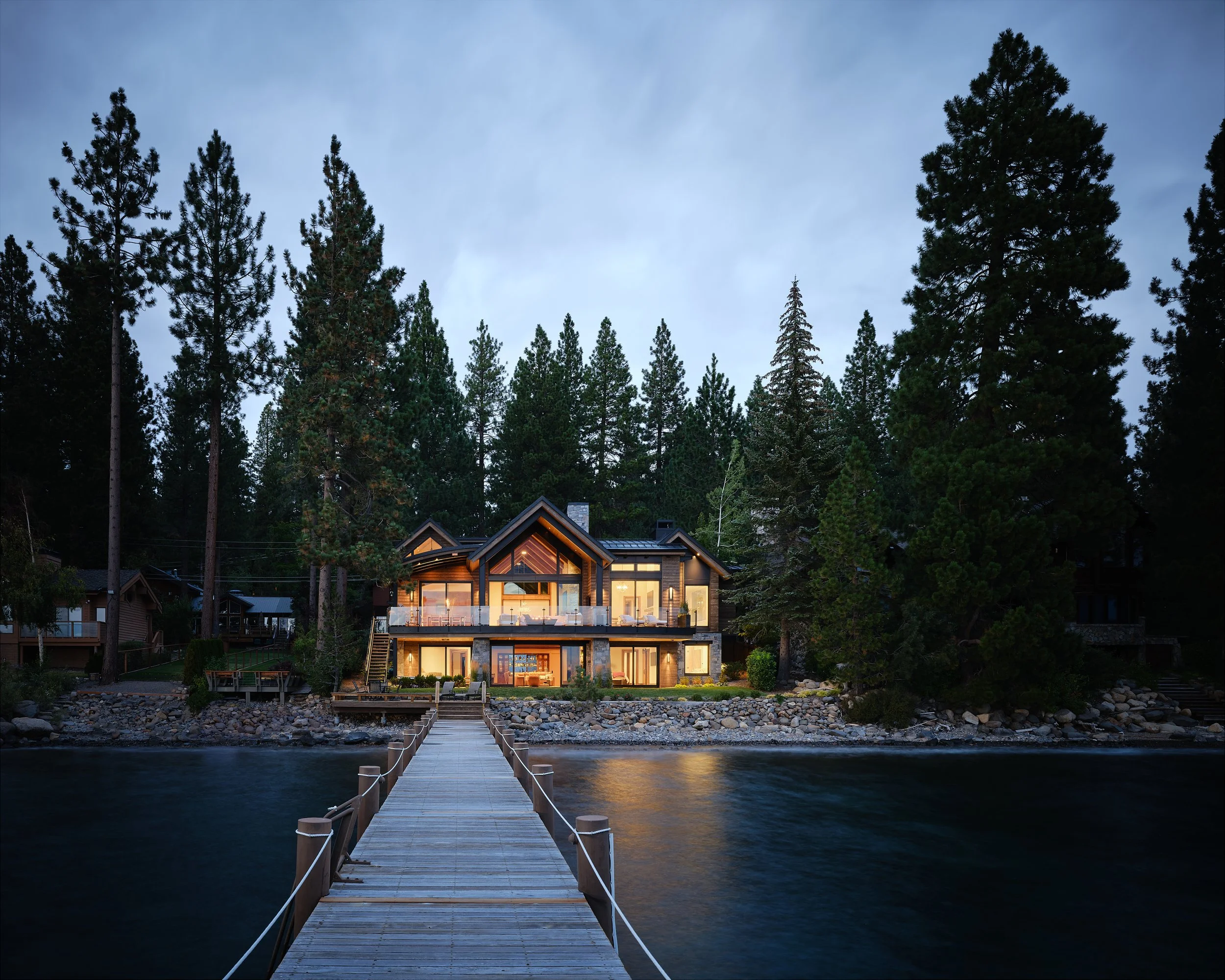 A modern house sits by a lake with a wooden dock leading to it. The house is illuminated from within, surrounded by tall pine trees, during dusk or evening.