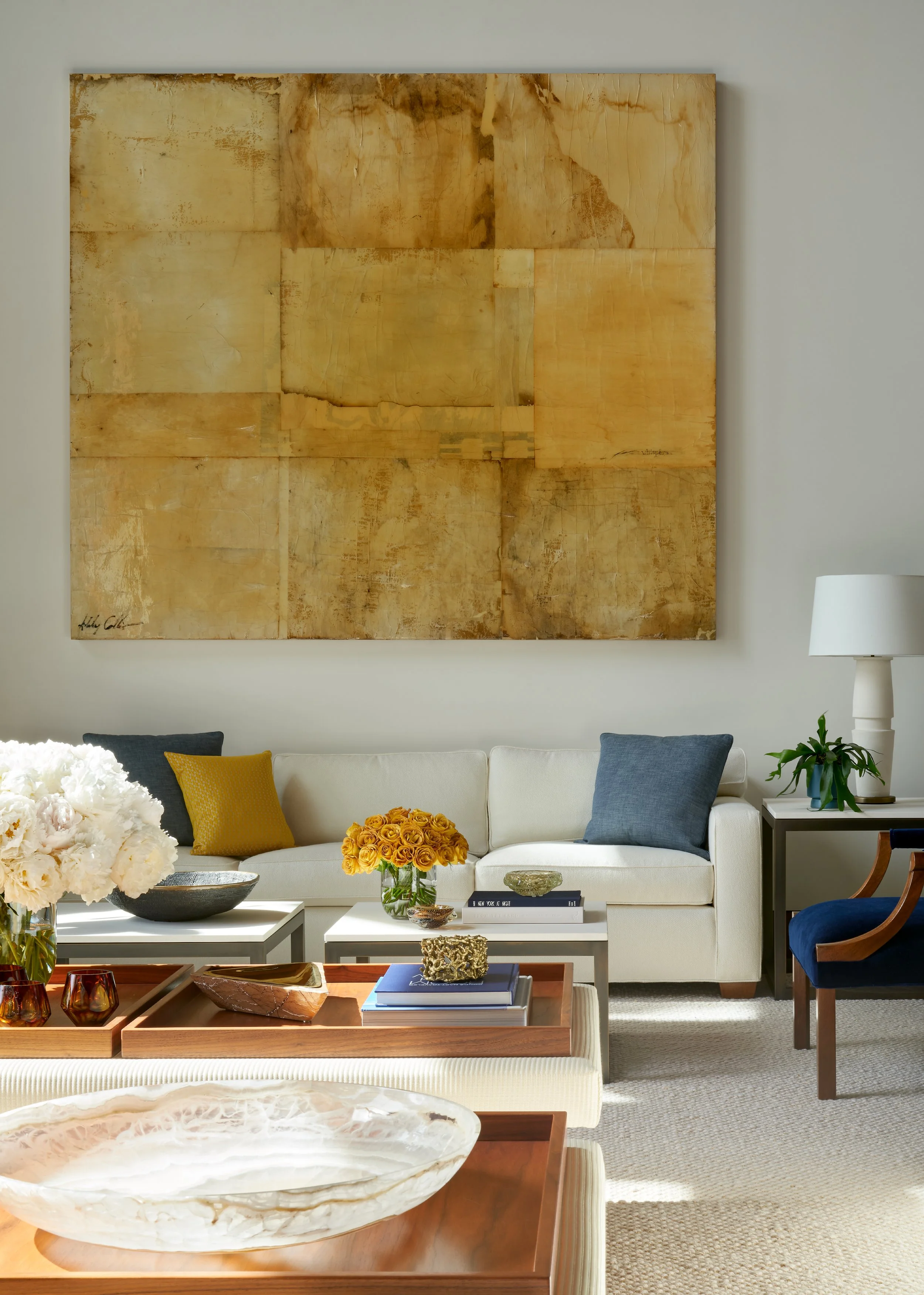 A modern living room with a white sofa, accent pillows, a large abstract gold artwork on the wall, a coffee table with decorative items, and a side table with a lamp and plant.