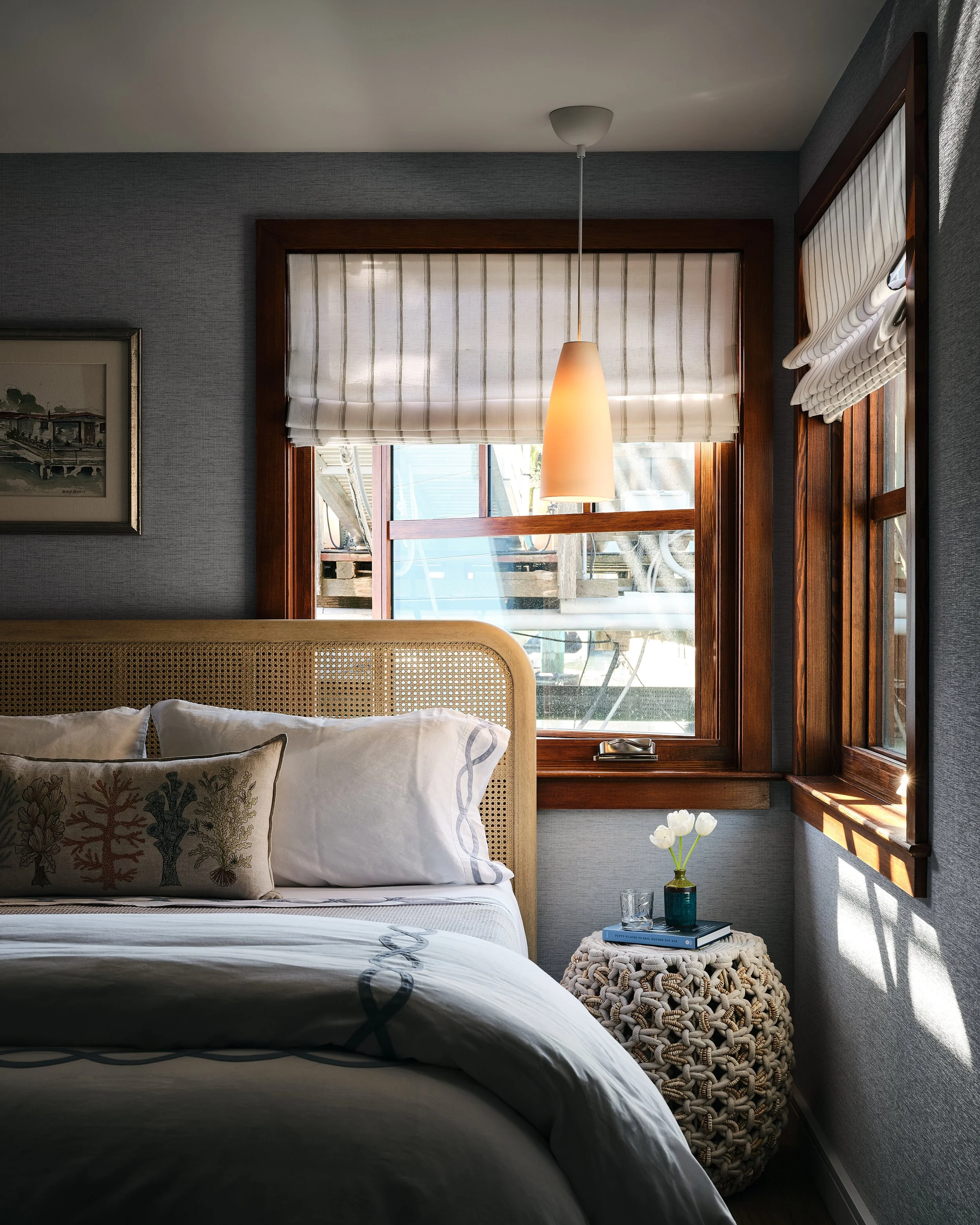A cozy bedroom with a bed, decorative pillows, a woven headboard, a small side table with a vase of white flowers, books, and glass, and two windows with wooden frames and striped Roman shades, lit by a hanging pendant light.