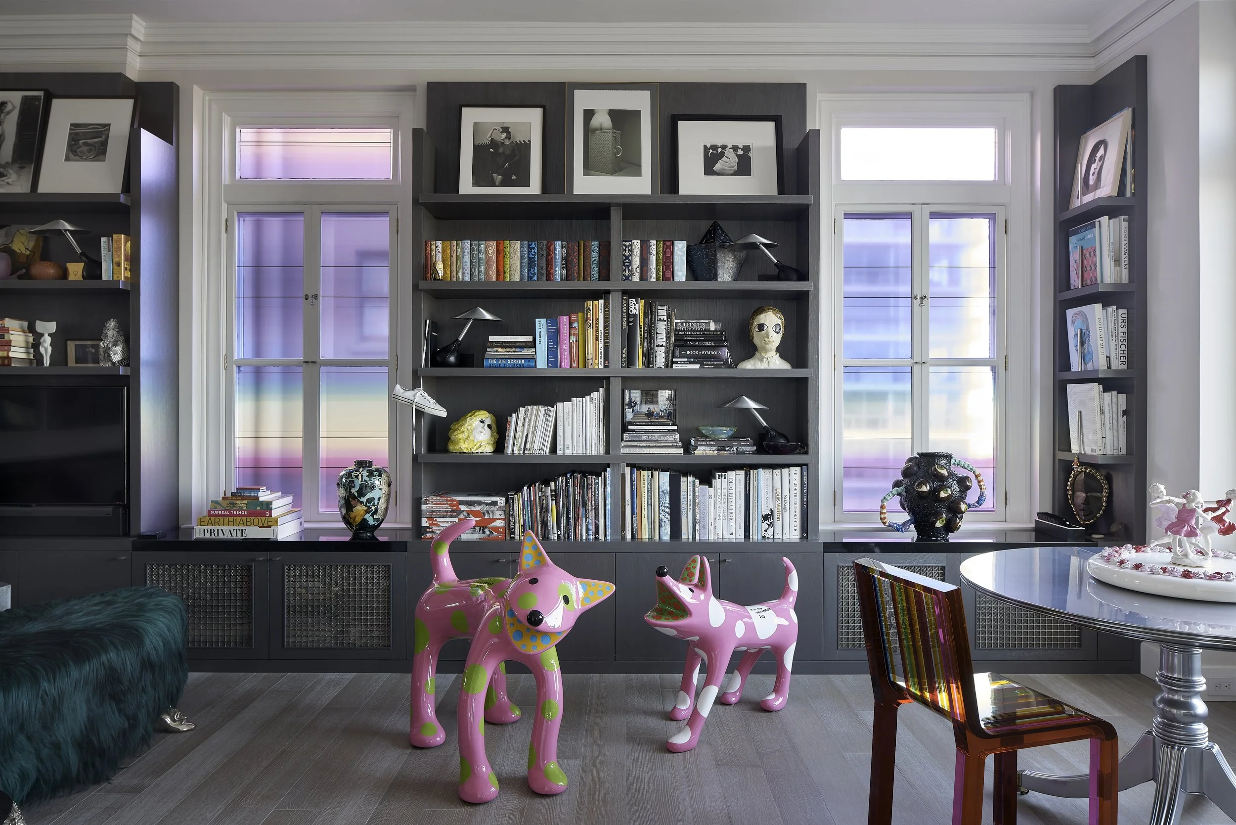 Living room with dark bookshelves filled with books and art, two large windows with colorful curtains, pink and green dog sculptures on the floor, and a round table with a decorative piece on top.