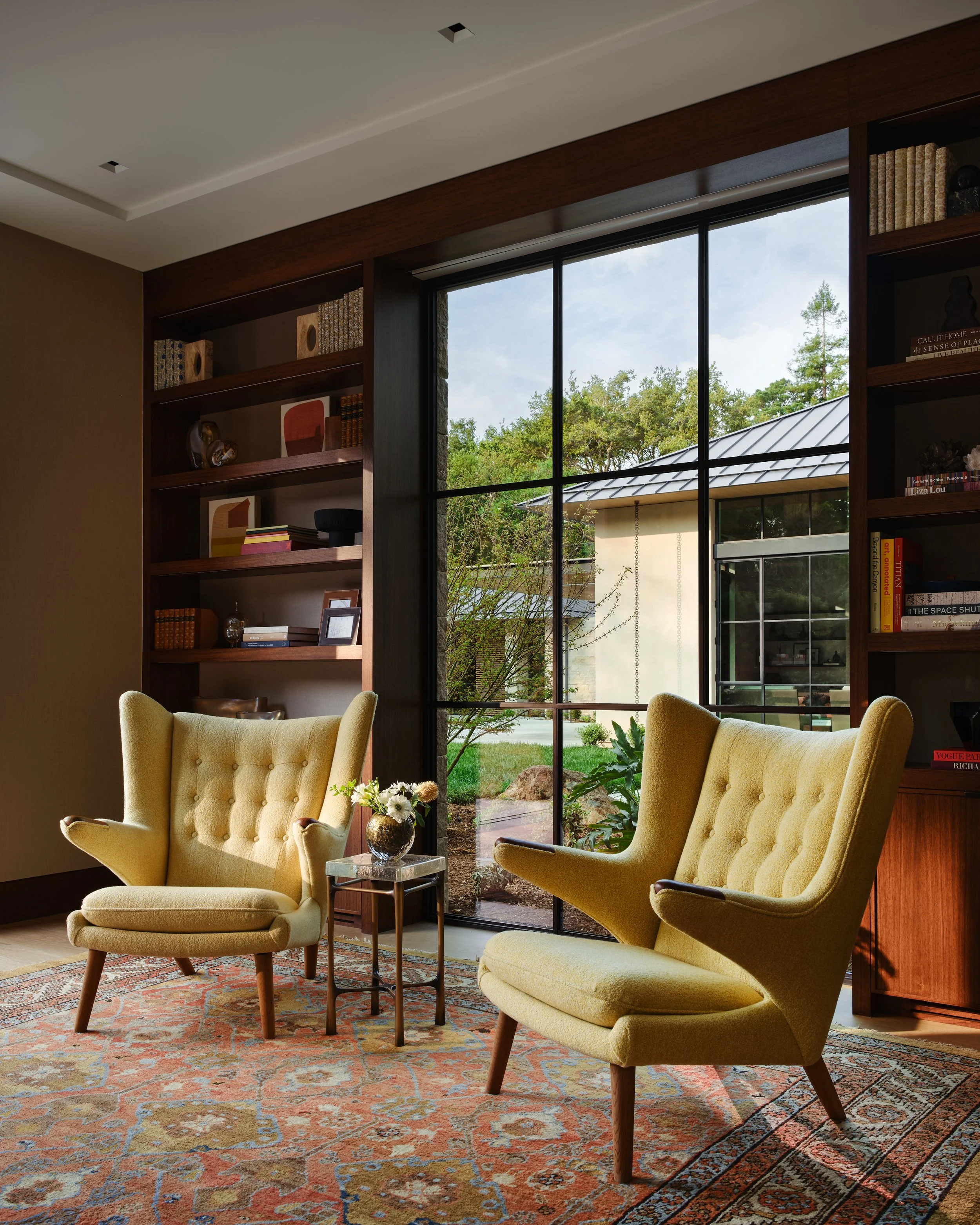 Living room with two yellow wingback chairs, a side table with a flower arrangement, a large window with black framing, bookshelves with books and decorative items, and a patterned area rug.Interior design and Architecture. California architectural p