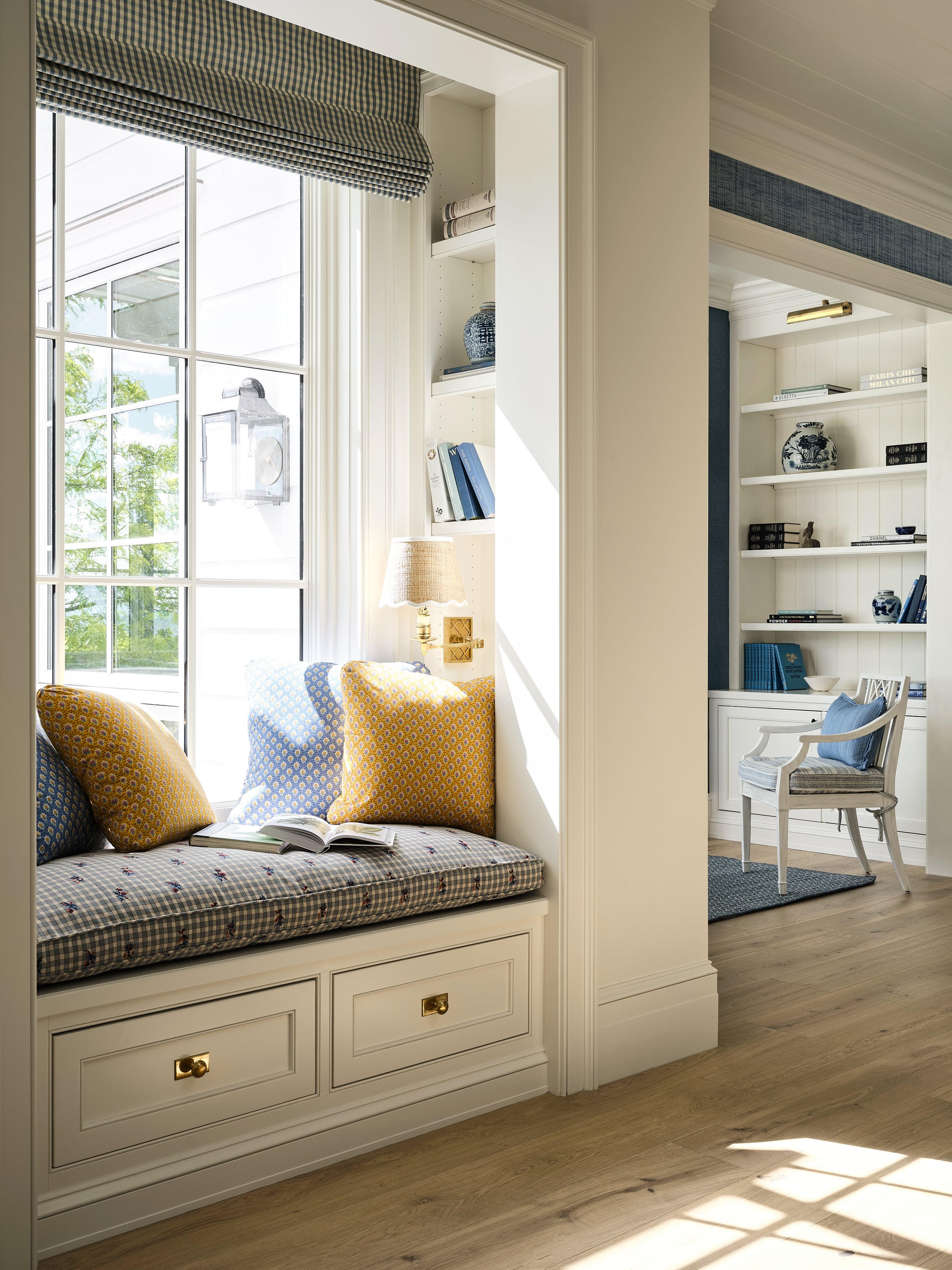 A cozy sunlit nook with a window seat featuring checkered and patterned cushions, a book open on the seat, next to a window with a checkered valance, and a built-in light fixture. The adjacent room has a white bookshelf with decorative vases and book