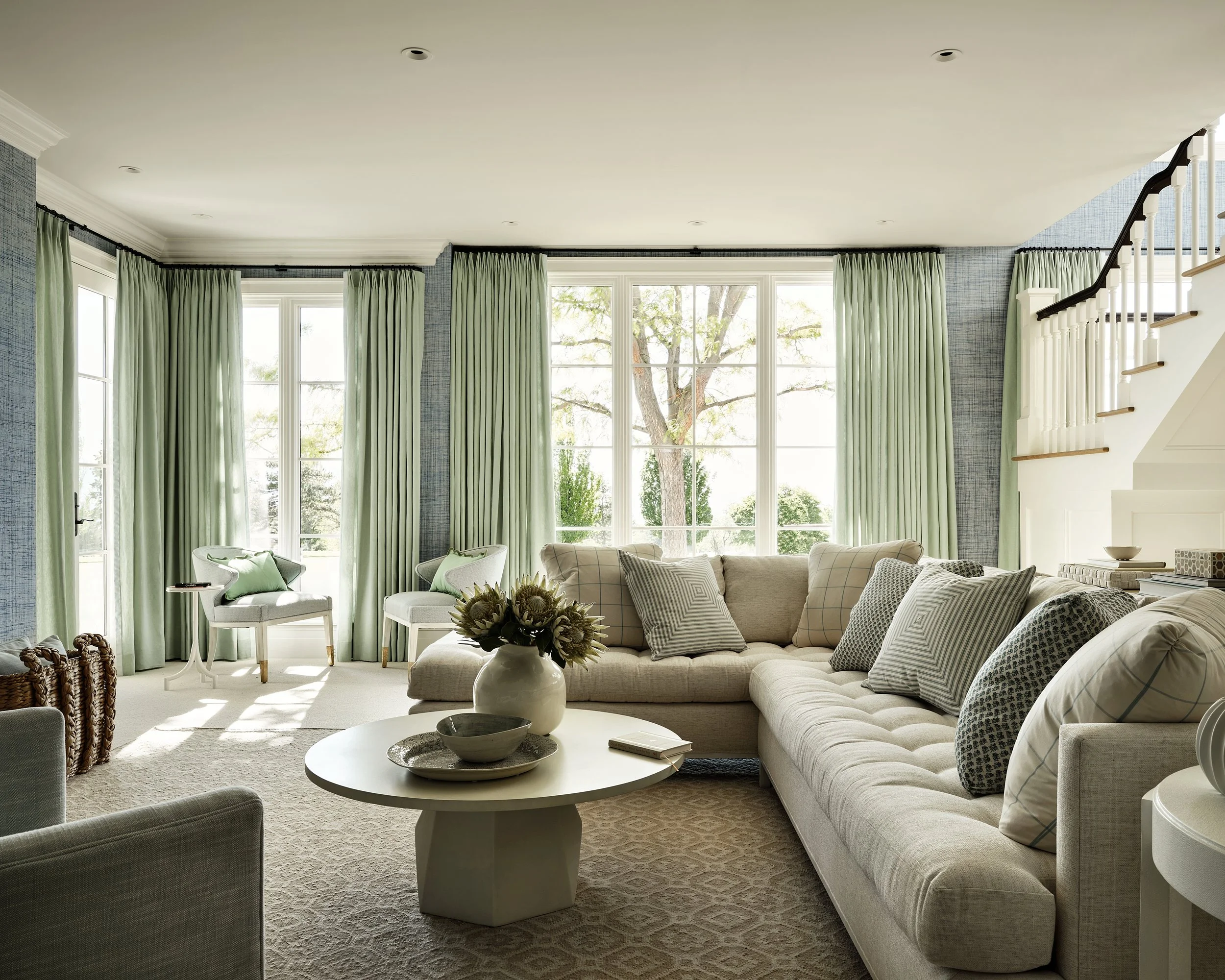 Bright living room with large windows, light green curtains, a sectional sofa with multiple pillows, a round coffee table with a vase of flowers, and stairs with white railing in the background.