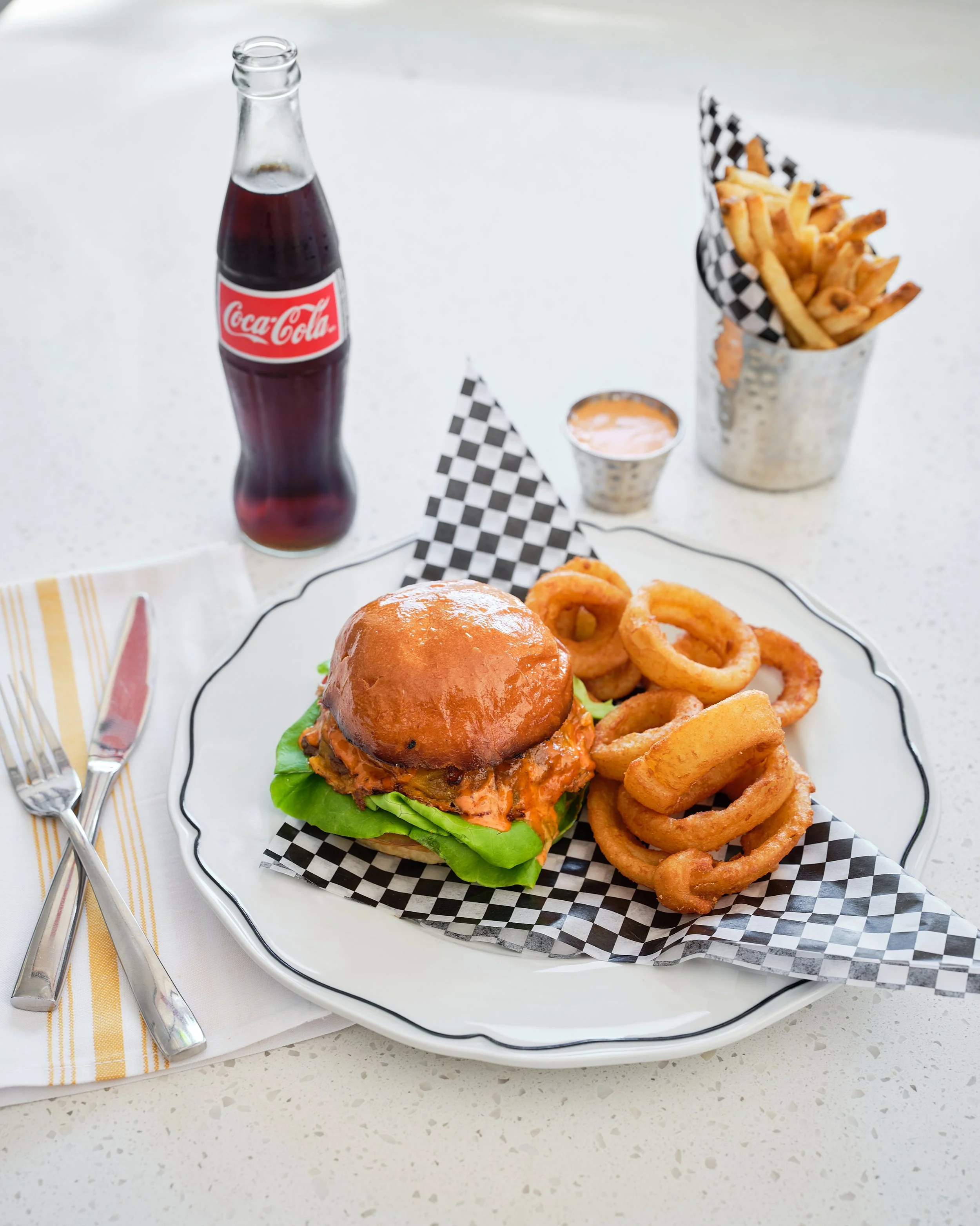 A plate with a burger, onion rings, and checkered paper, accompanied by a bottle of Coca-Cola, a small cup of dipping sauce, and a container of French fries, all on a white table.