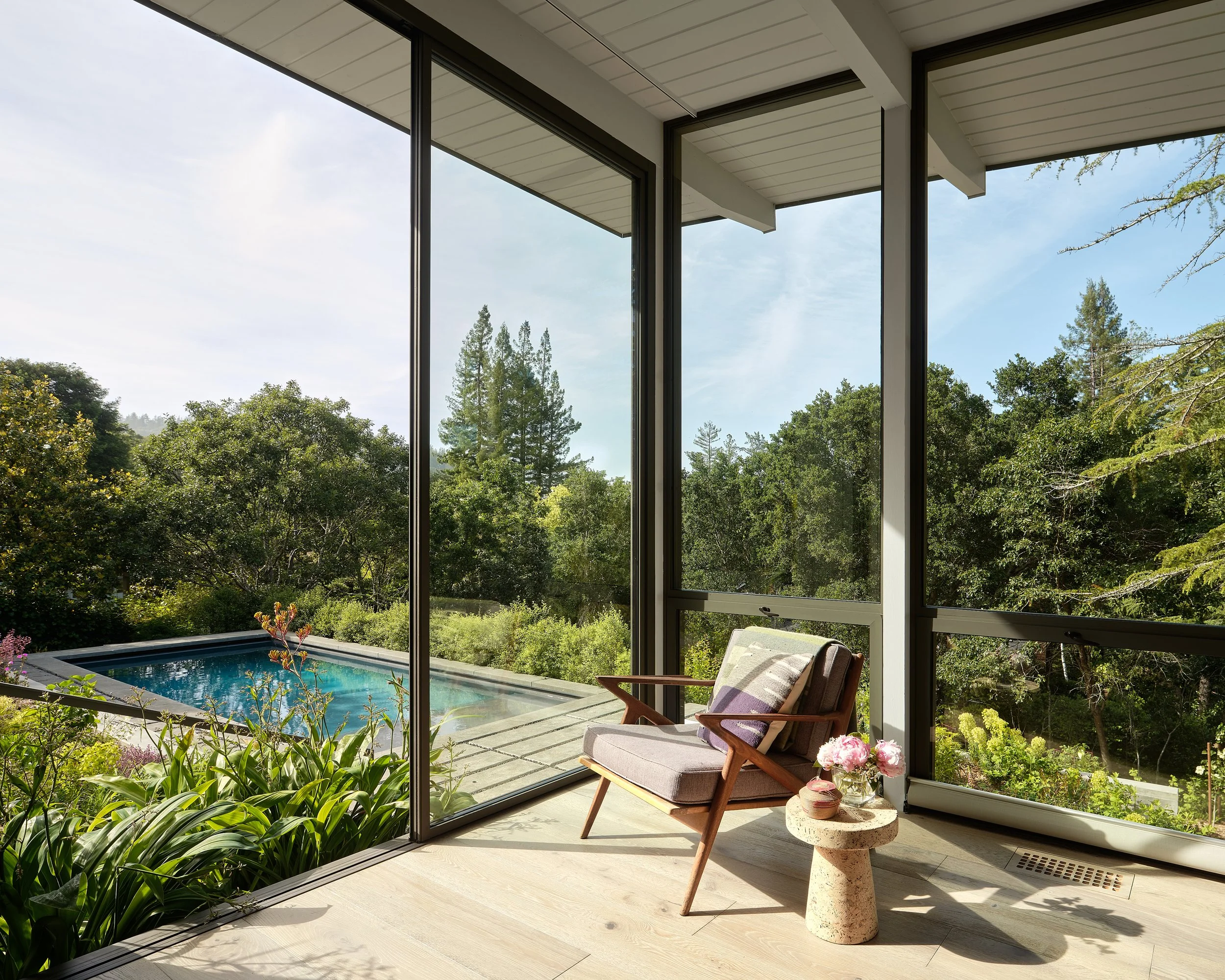 Sunlit room with large glass windows facing a lush green landscape and a backyard with a small swimming pool; contains a mid-century modern chair with a cushion, a small stone side table with pink flowers, and a floor vent.