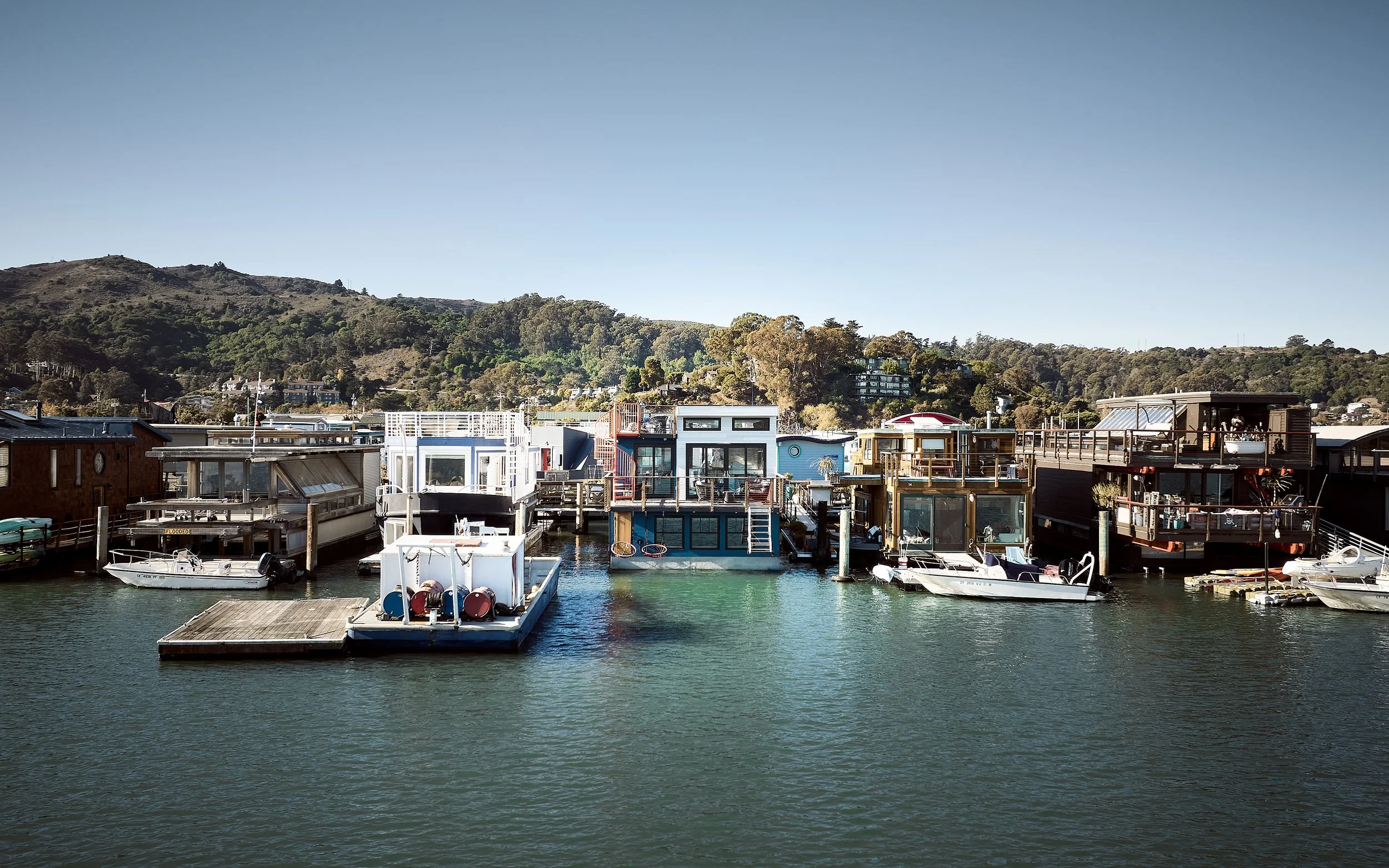 View of houseboats docked along a waterfront with watercraft and boats, hillside with trees and buildings in the background, and clear blue sky.