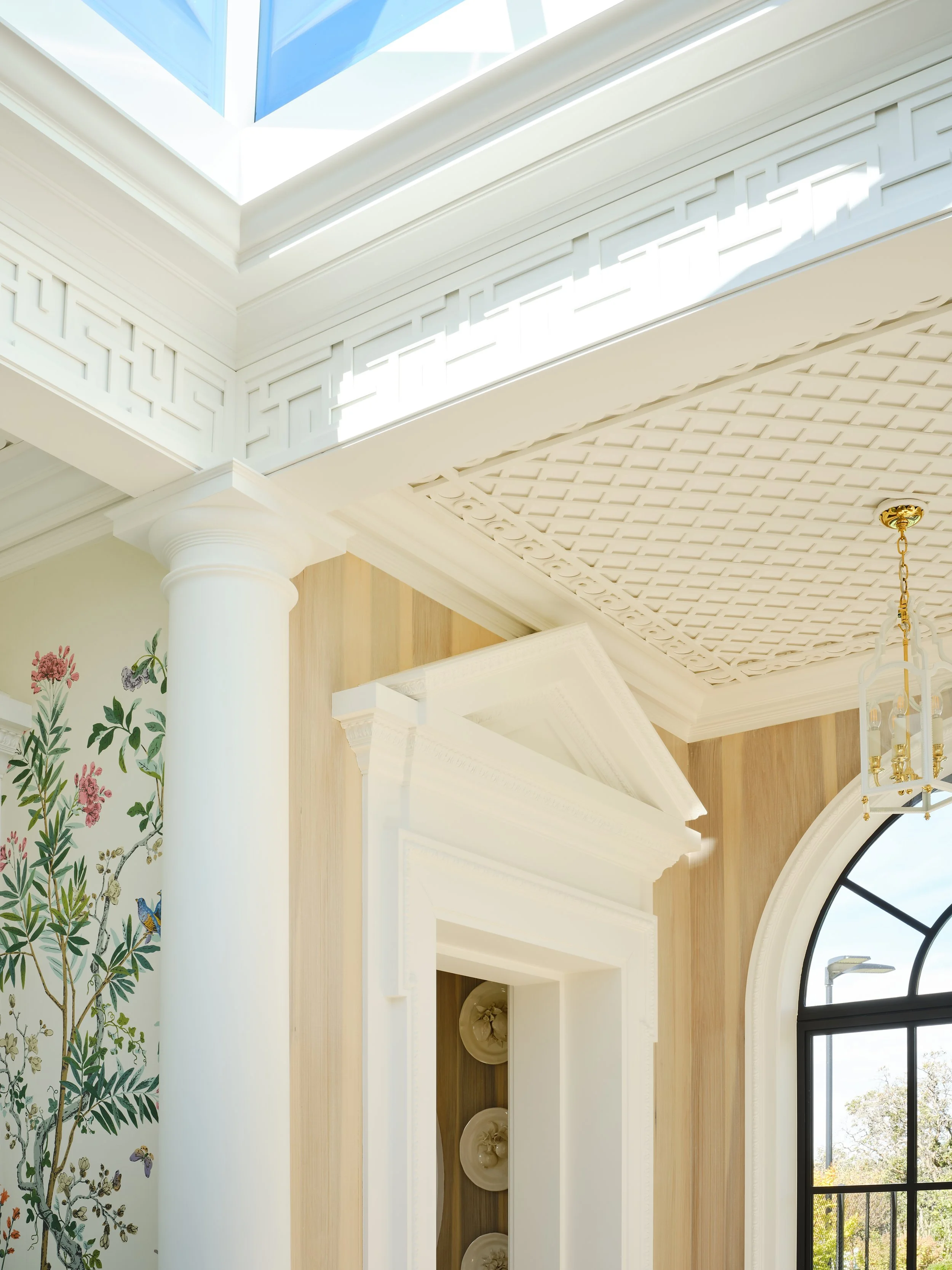 Interior view of a room with a large arched window, white columns, decorative crown molding, a painted floral wall, and a ceiling with a lattice pattern and a hanging chandelier with gold accents.