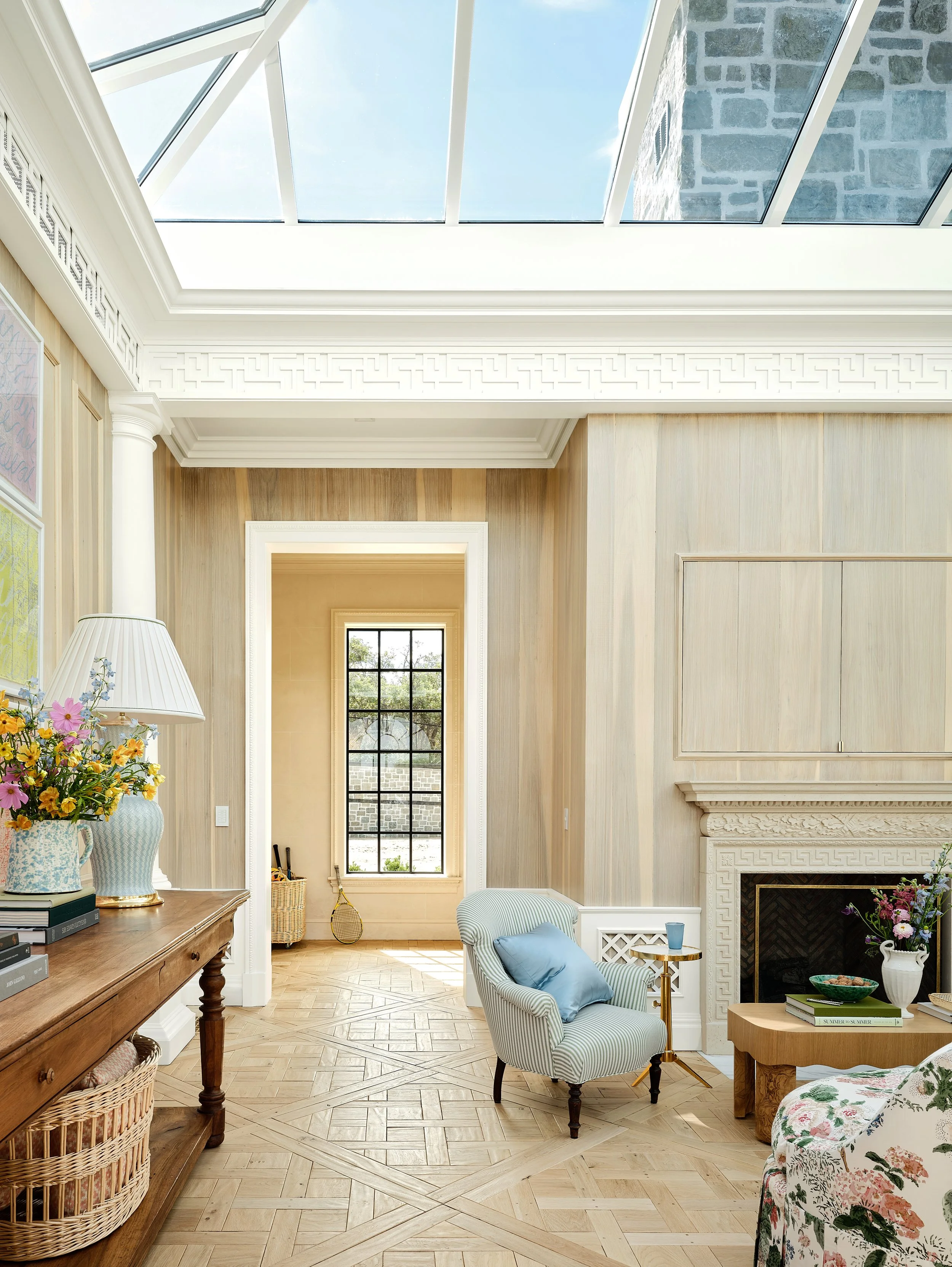 A bright living room with wood-paneled walls, a large window at the back, a fireplace, and a skylight ceiling allowing natural light. Features include a striped armchair with a blue pillow, a wooden table with books and a vase of flowers, and decorat