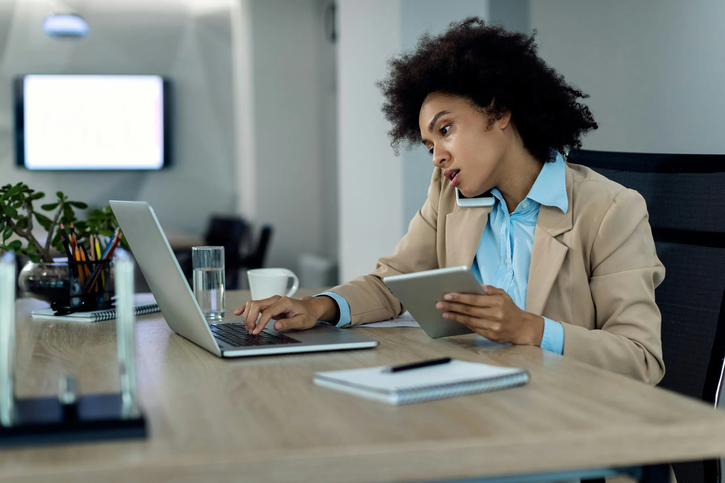 a woman talks on the phone while holding a tablet and working on a laptop