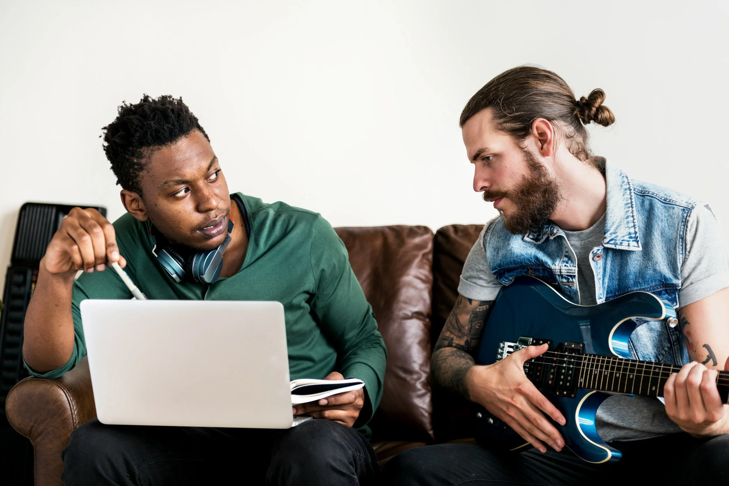 one man works on a laptop next to another man holding an electric guitar