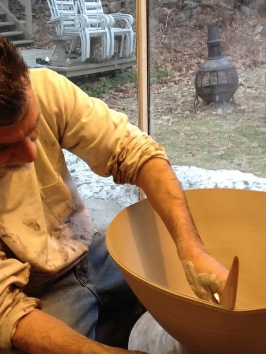 A man shaping a ceramic bowl on a pottery wheel inside a house, with a glass door revealing an outdoor area with stacked chairs and a chiminea.