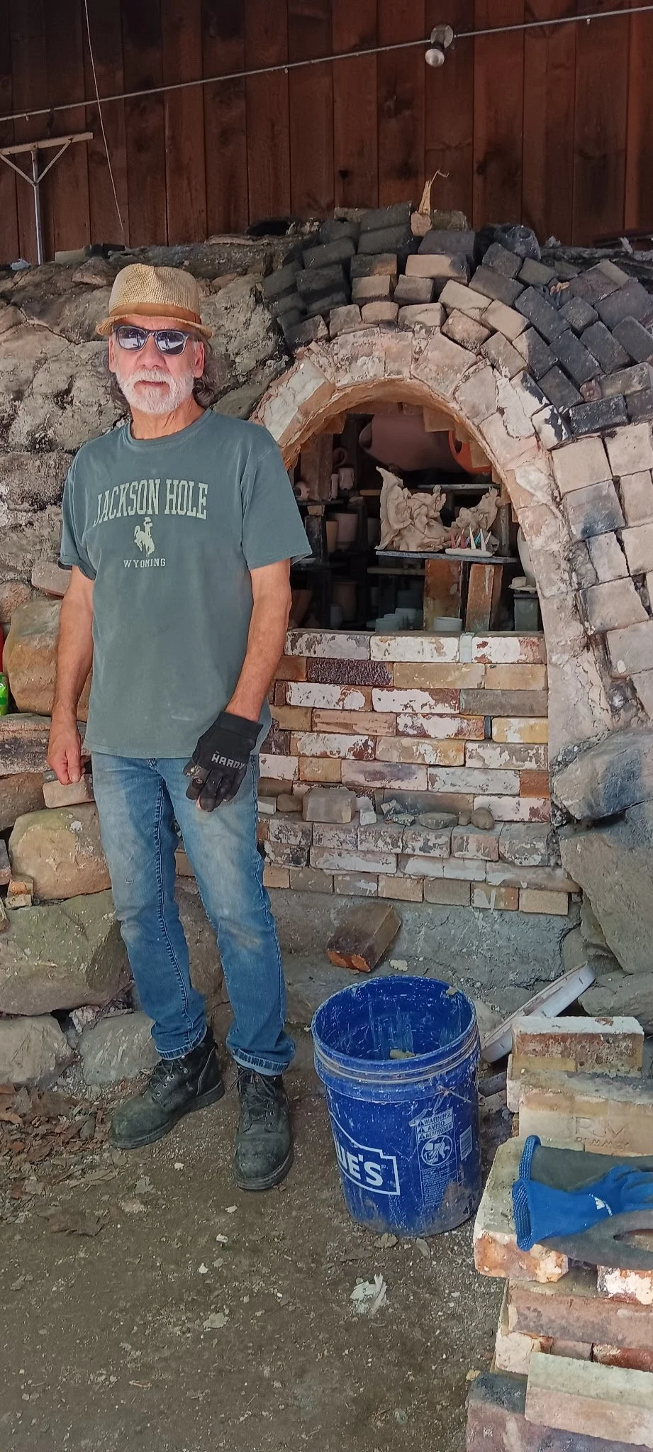 A man with sunglasses, a hat, gray beard, gray t-shirt that says 'Jackson Hole Wyoming,' jeans, gloves, and work boots stands in front of a partially constructed brick and stone fireplace.