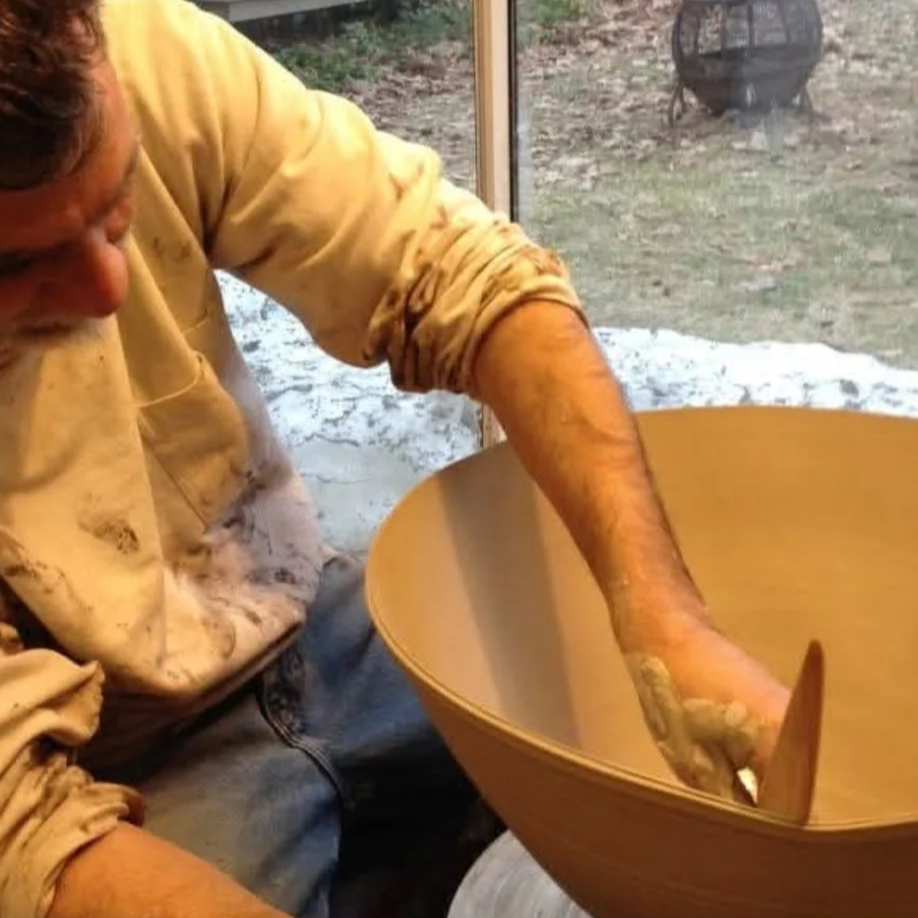 A man crafting a wooden bowl at a workshop, with a window showing an outdoor area with some snow.