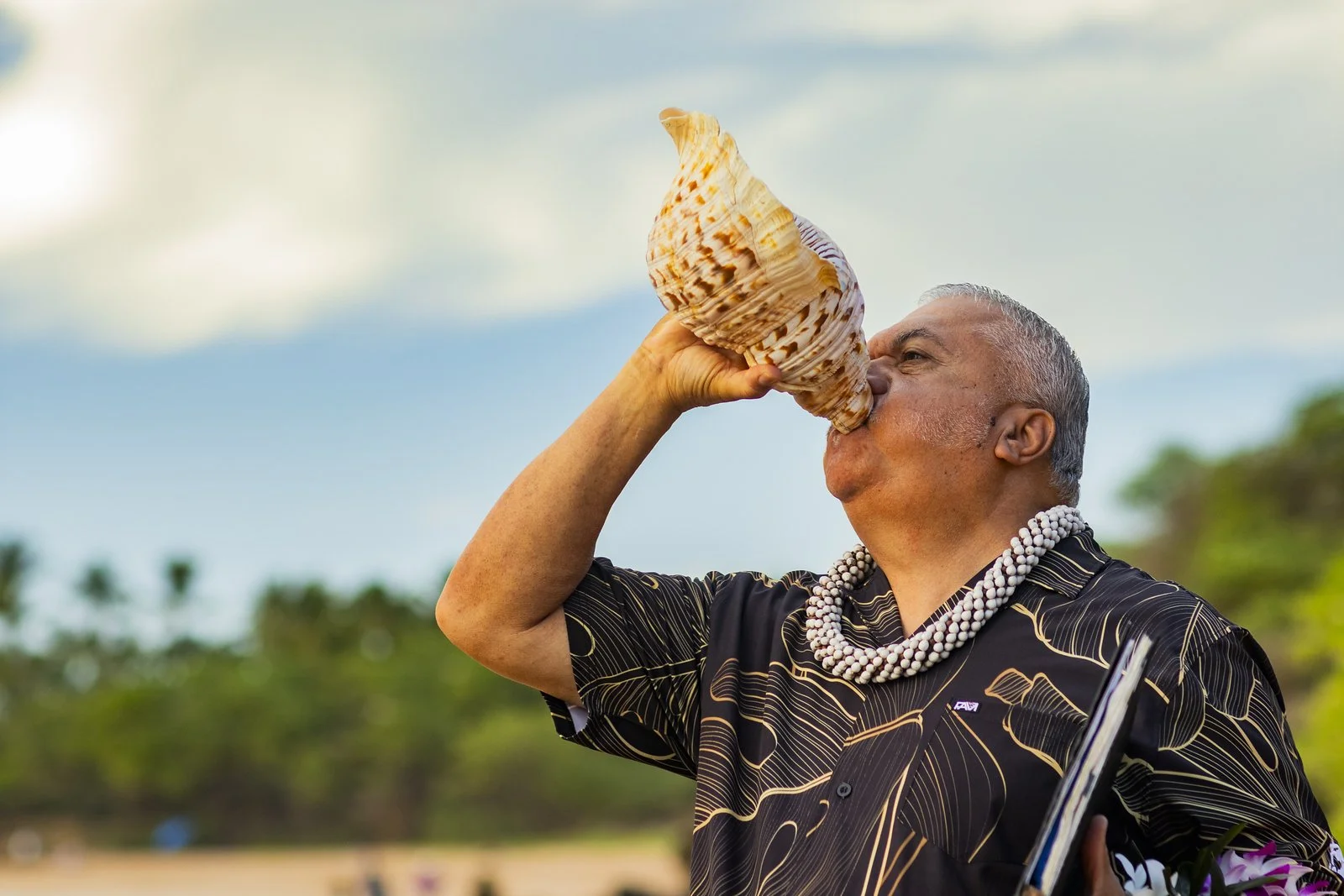 An elderly man with gray hair wearing a black Hawaiian shirt and a beaded necklace is drinking from a large seashell outdoors, with a background of trees and a cloudy sky.