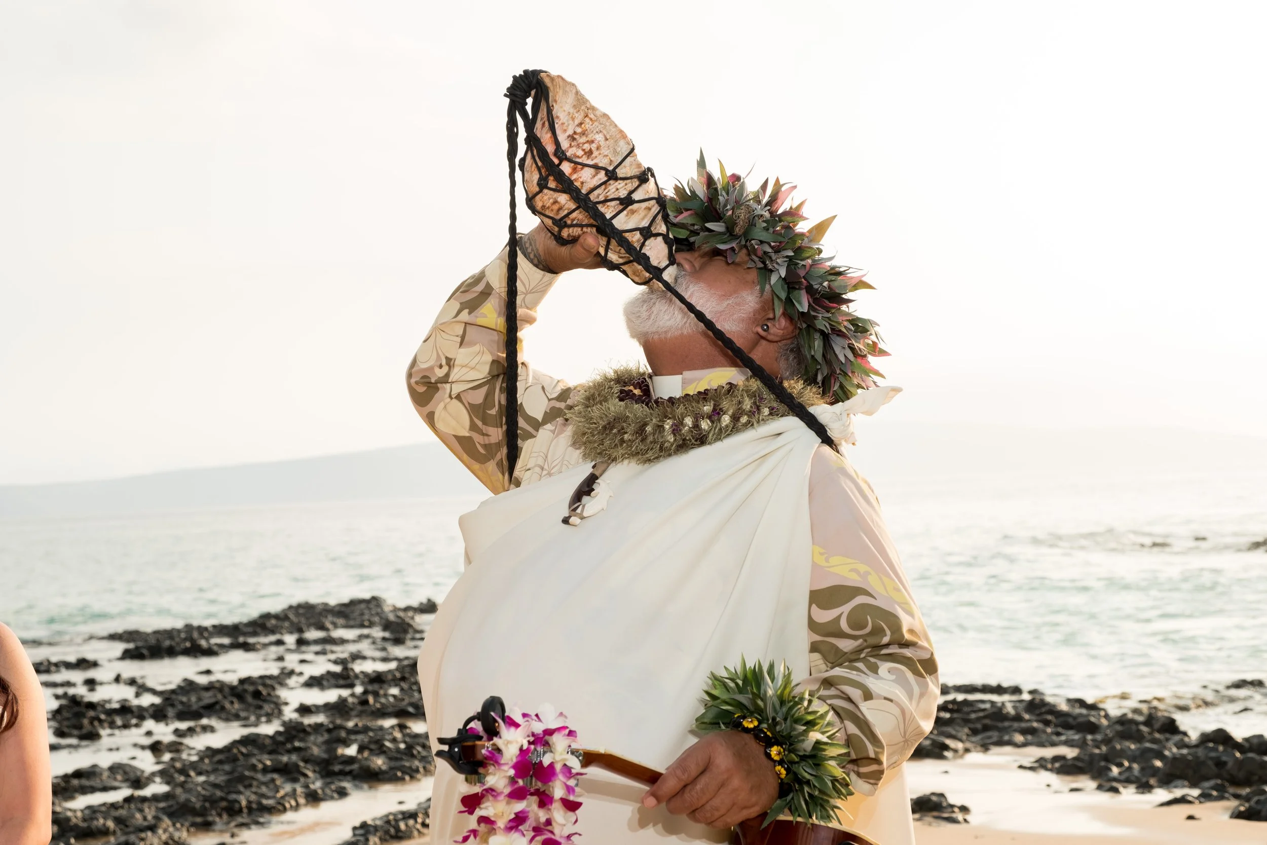 A man dressed in ceremonial attire, wearing a floral crown, and playing a shell horn on a rocky beach near the ocean.
