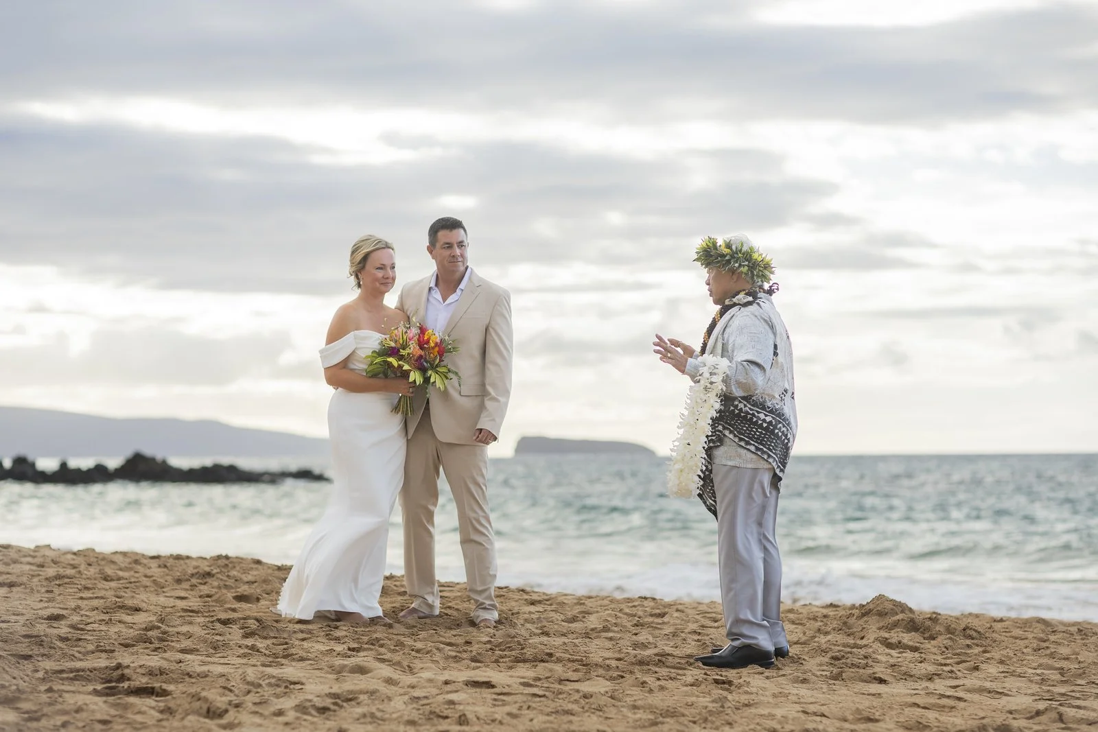 A couple getting married on the beach, standing with an officiant, with ocean waves and a cloudy sky in the background.
