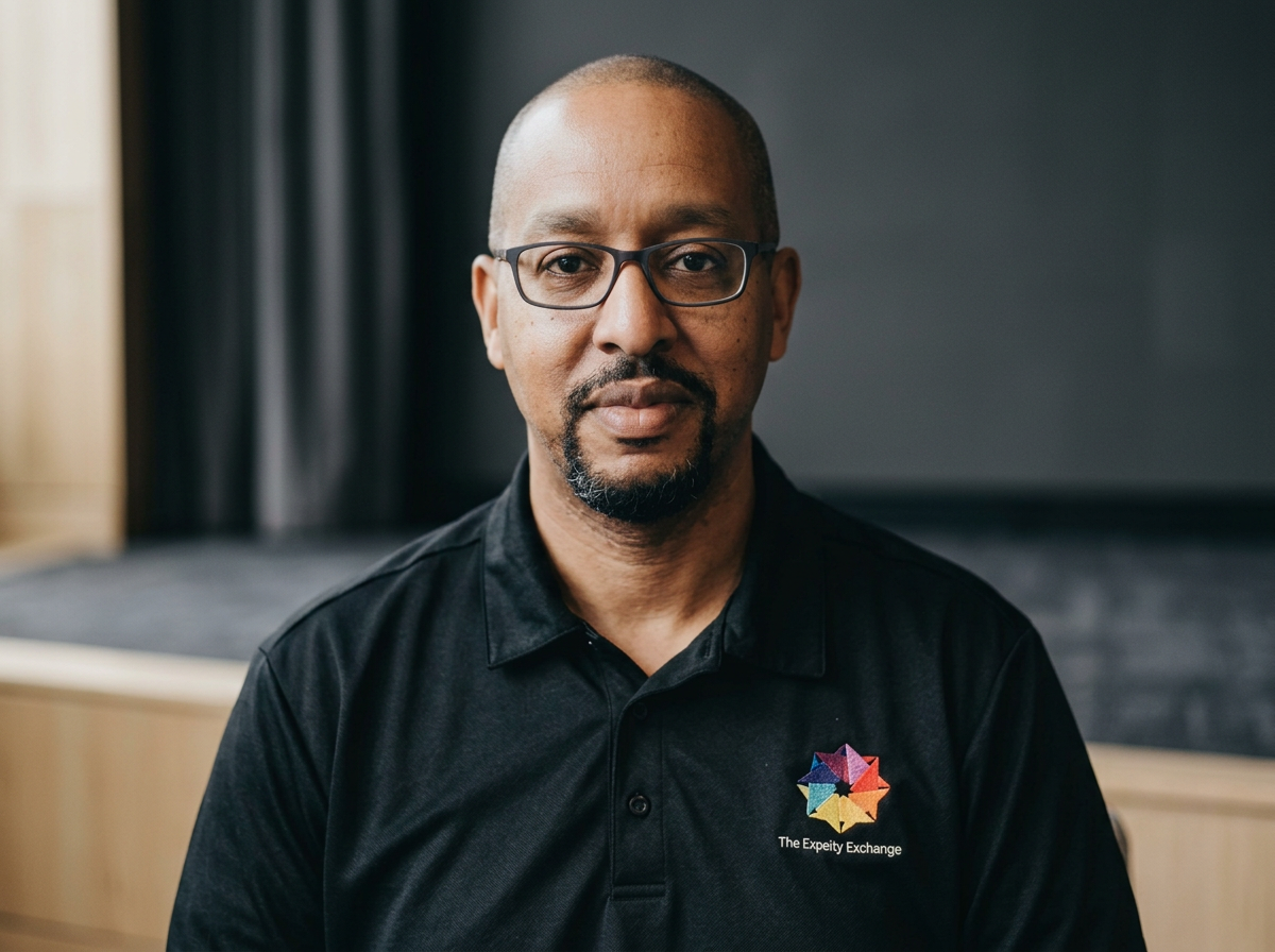 A man with glasses, a goatee, and a shaved head wearing a black polo shirt with a colorful logo and the text 'The Experty Exchange,' standing in a room with dark curtains and a blurred background.