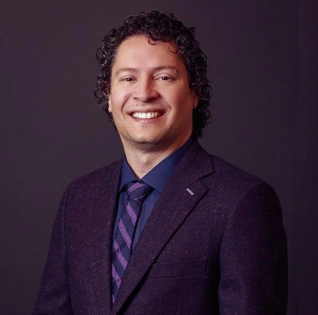 A man with curly hair smiling, wearing a dark blazer, blue shirt, and a striped purple and blue tie against a dark background.