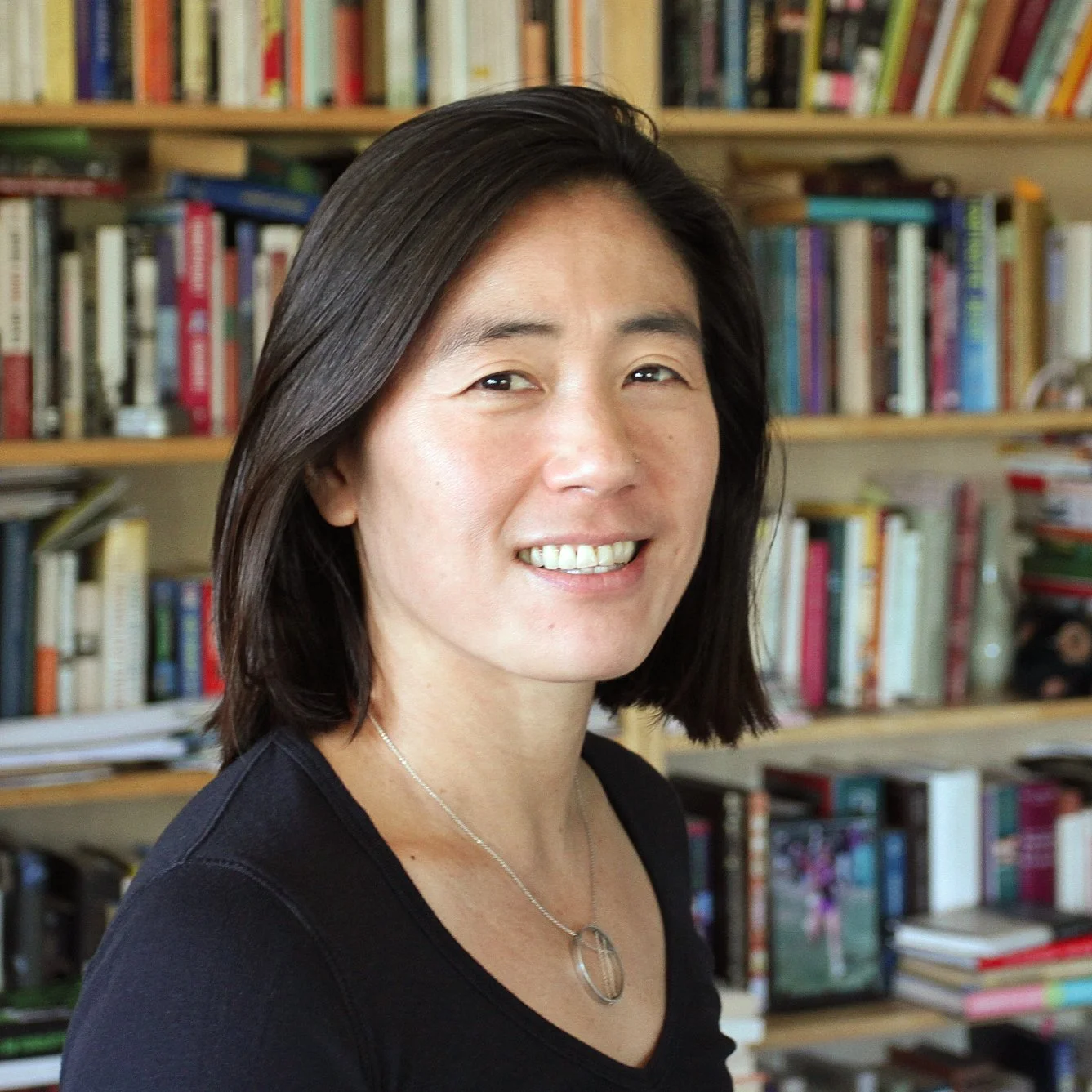 A woman with shoulder-length dark hair smiling in front of a bookshelf filled with colorful books.