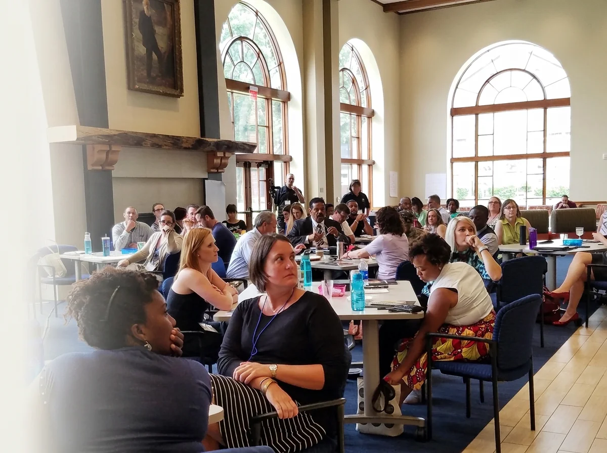 A diverse group of people attending a conference or workshop in a bright, spacious room with large arched windows. They are seated at tables, listening attentively.