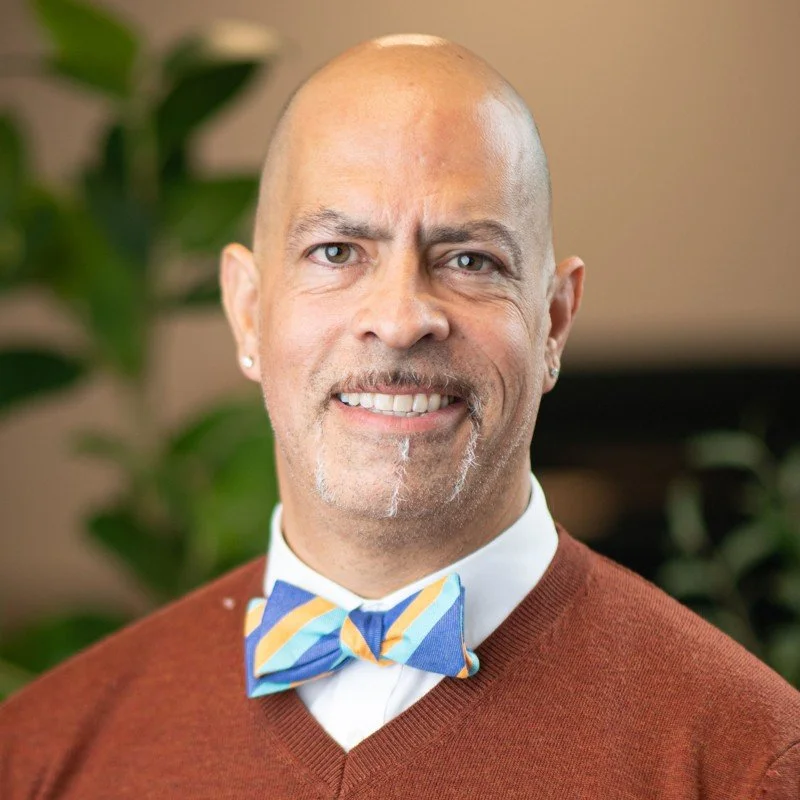 Close-up portrait of a bald man with light skin, smiling, wearing a colorful striped bow tie and a brown sweater, with a green plant in the background.