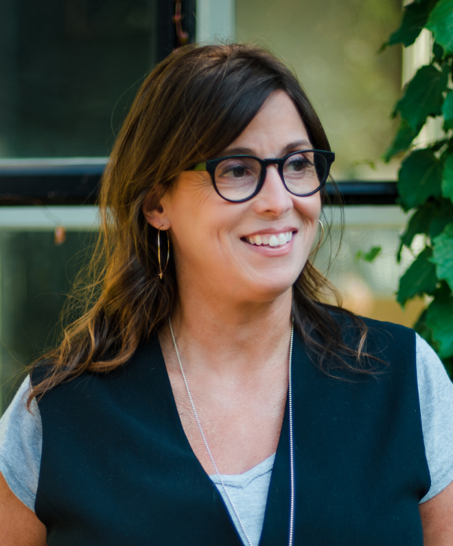 A woman with brown hair, wearing glasses, a black vest over a gray shirt, and earrings, smiling indoors near green plants.