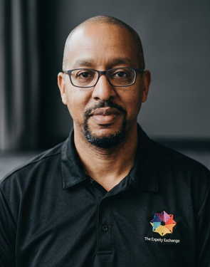 Portrait of a man wearing glasses and a black polo shirt with a colorful logo, indoors with a dark background.