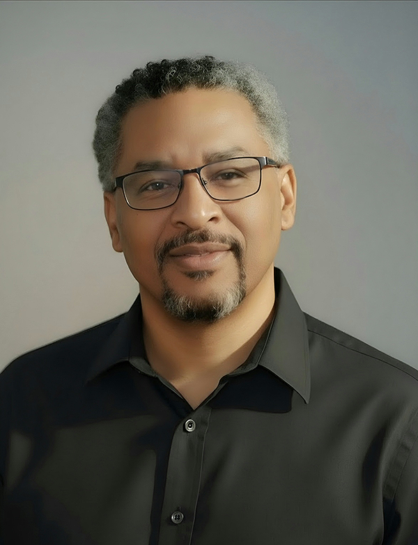 A man with glasses and a beard wearing a black collared shirt, smiling softly against a gray background.