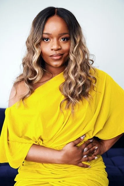 Woman with wavy hair and a yellow dress, standing with one hand on her waist against a plain background.