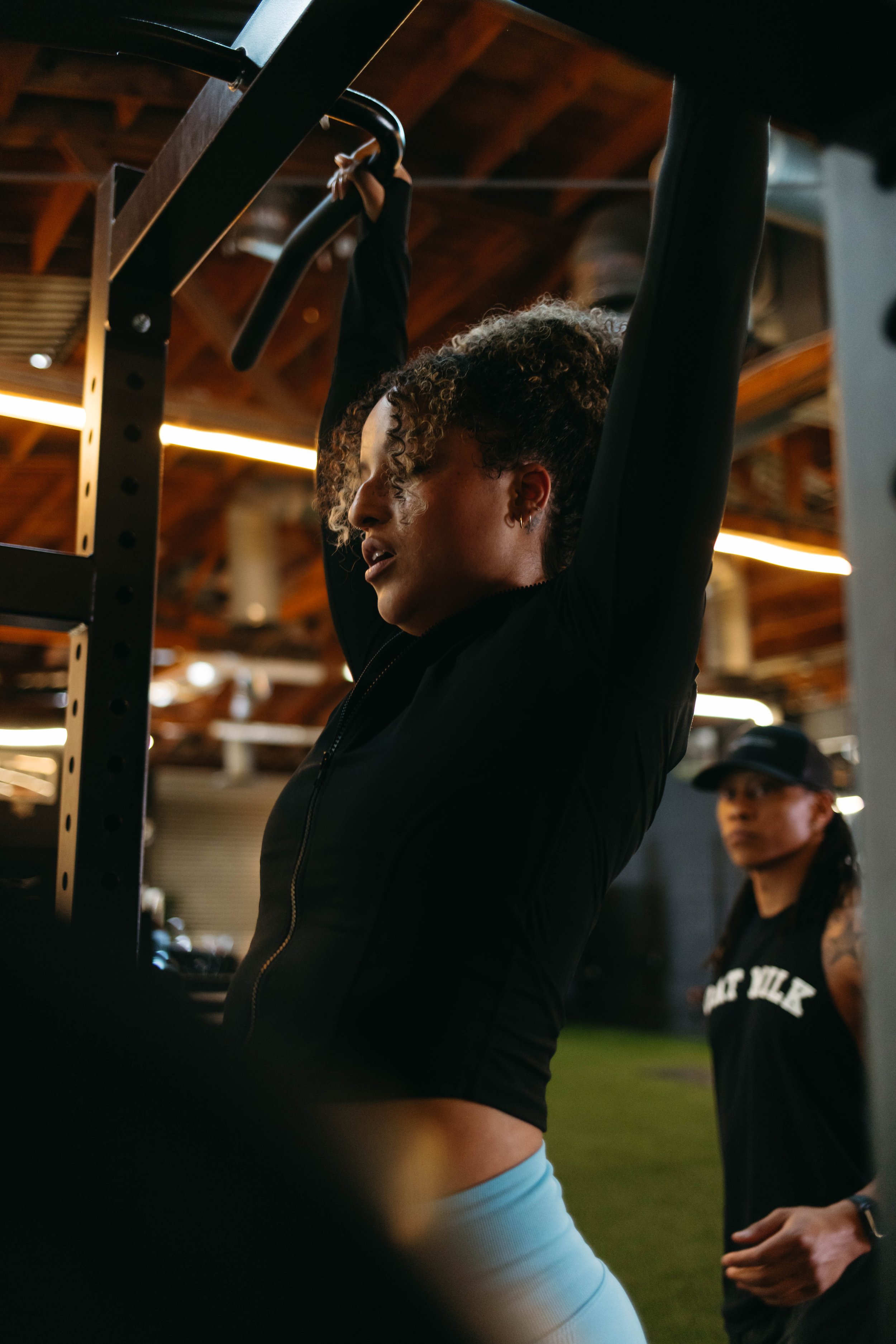 Woman doing pull-ups in a gym with another person watching, wearing a black jacket and light-colored pants, under a wood-paneled ceiling.