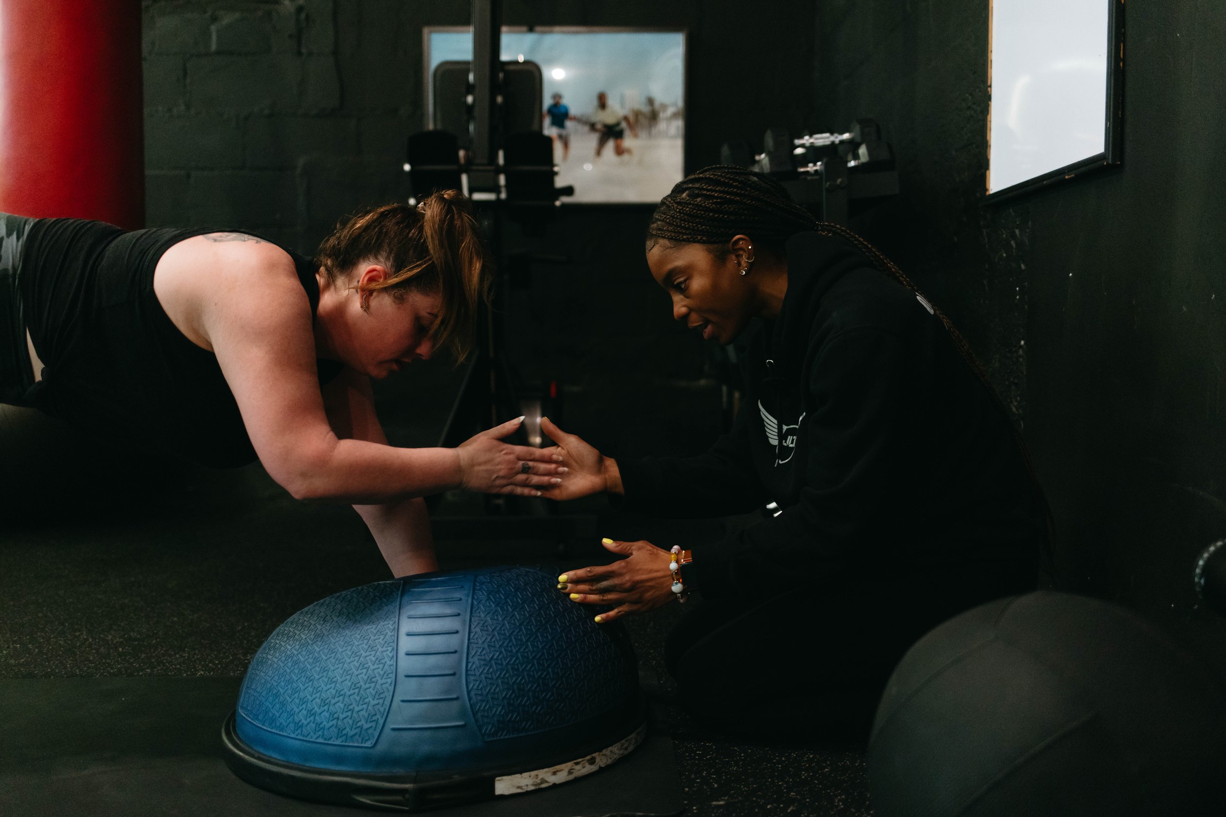 A woman engaging in exercise with a trainer, using a blue Bosu balance trainer. The scene is set in a gym with dark walls and fitness equipment in the background. The trainer is crouched, assisting or encouraging her.