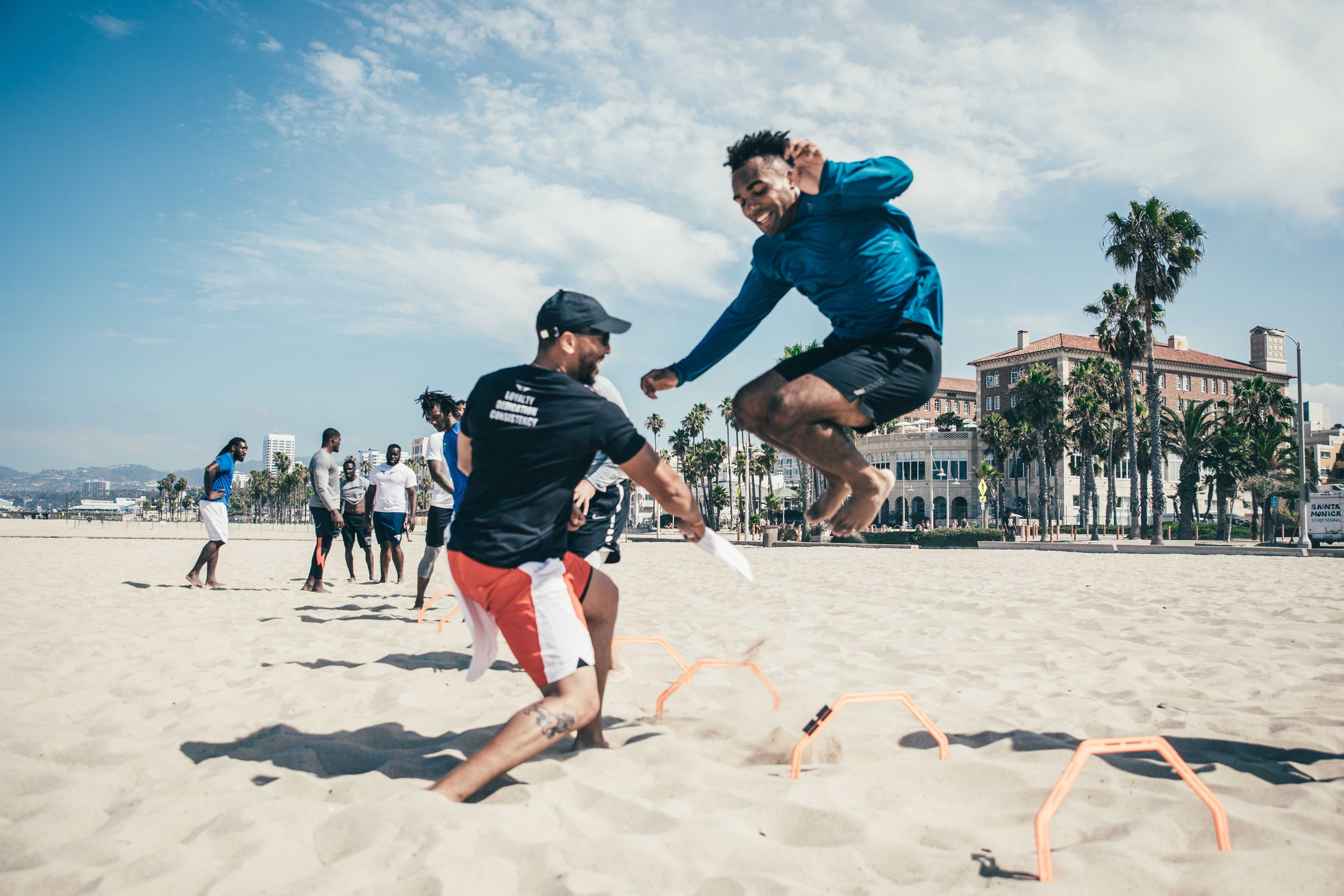 Group of people training on Santa Monica Beach, performing agility drills on sand. One person is jumping over hurdles, while others observe under a clear sky. Palm trees and buildings are visible in the background. christian Kirk