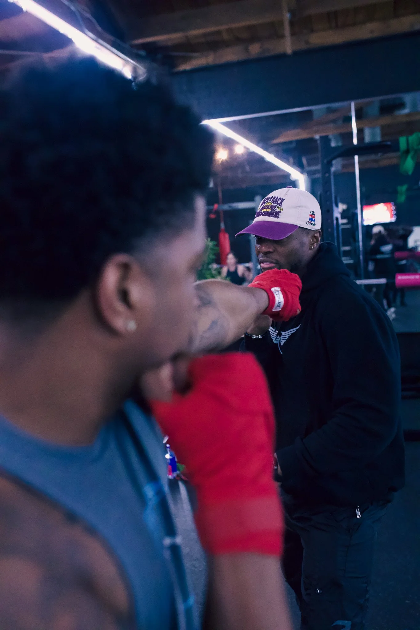 Two men practicing boxing in a gym, one wearing red hand wraps and the other a white and purple cap, with a punching bag visible in the background.